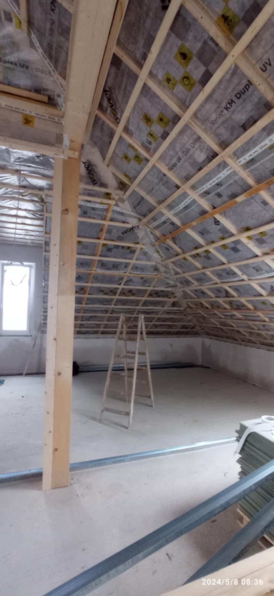 Poddasze w trakcie ocieplania: widoczne drewniane krokwie, folia paroizolacyjna z nadrukiem Isover, drabina i profile. Prace wykończeniowe w surowym stanie.