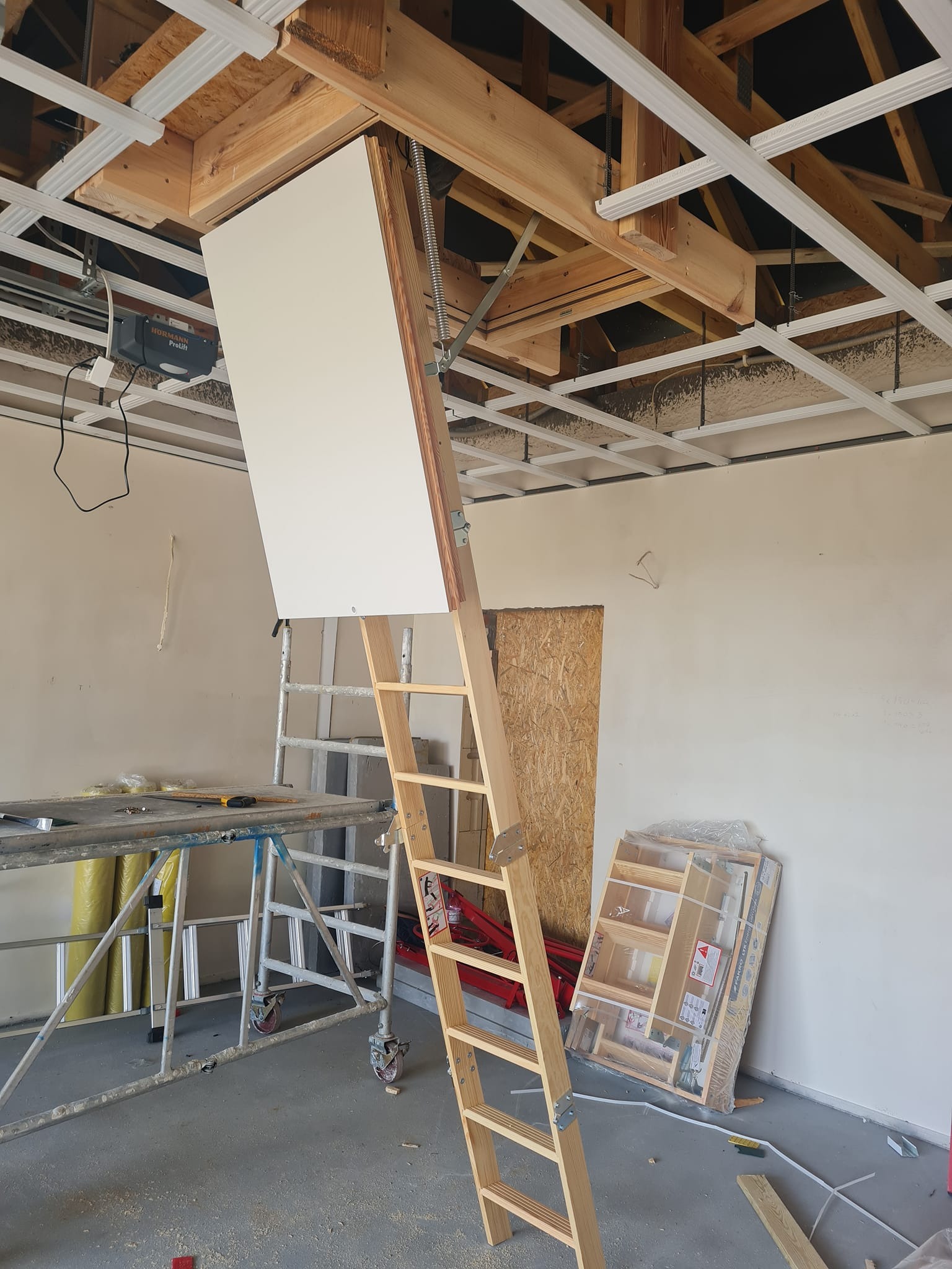Montaż drewnianych schodów strychowych z białą klapą w pomieszczeniu z częściowo zamontowanym rusztem sufitowym i narzędziami pracy w tle.