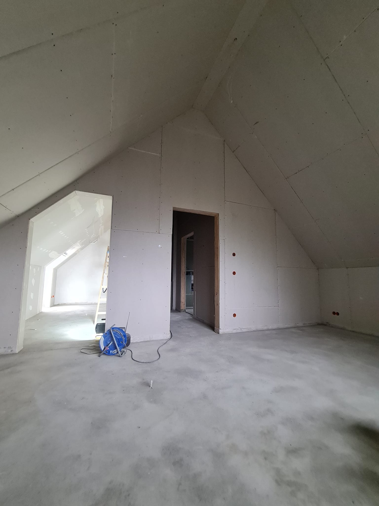 Poddasze z zamontowanymi płytami gipsowo-kartonowymi na ścianach i skosach. Widoczne otwory drzwiowe i narzędzia. Surowy stan wykończenia, brak szpachlowania.