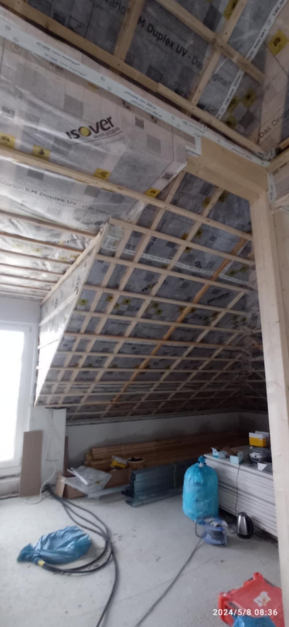 Poddasze w trakcie ocieplania z widoczną konstrukcją drewnianą, folią izolacyjną Isover i materiałami budowlanymi. Prace ociepleniowe w toku.