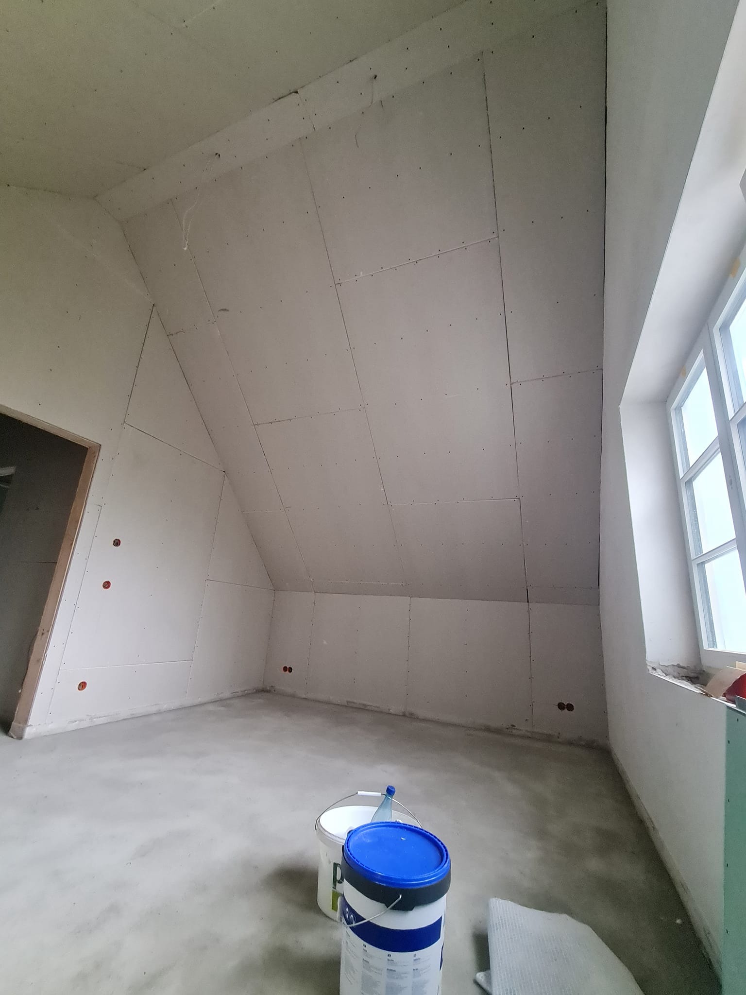 Poddasze z oknem po montażu płyt kartonowo-gipsowych na skosach i ścianach, widoczne puszki elektryczne. Na podłodze wiaderka z farbą.