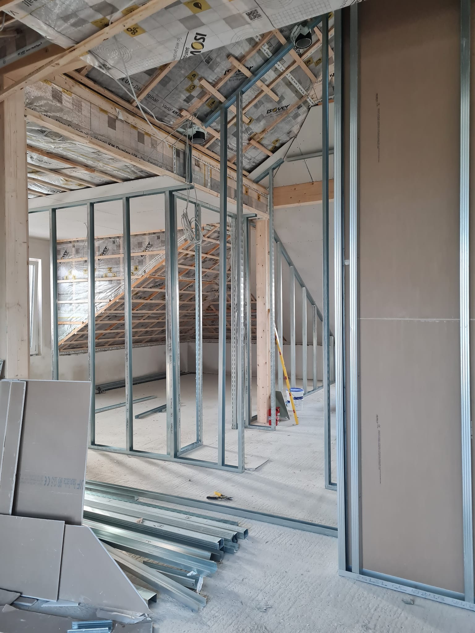 Montaż metalowej konstrukcji ścian działowych w surowym wnętrzu poddasza z izolacją termiczną. Widoczne profile, płyty i narzędzia.