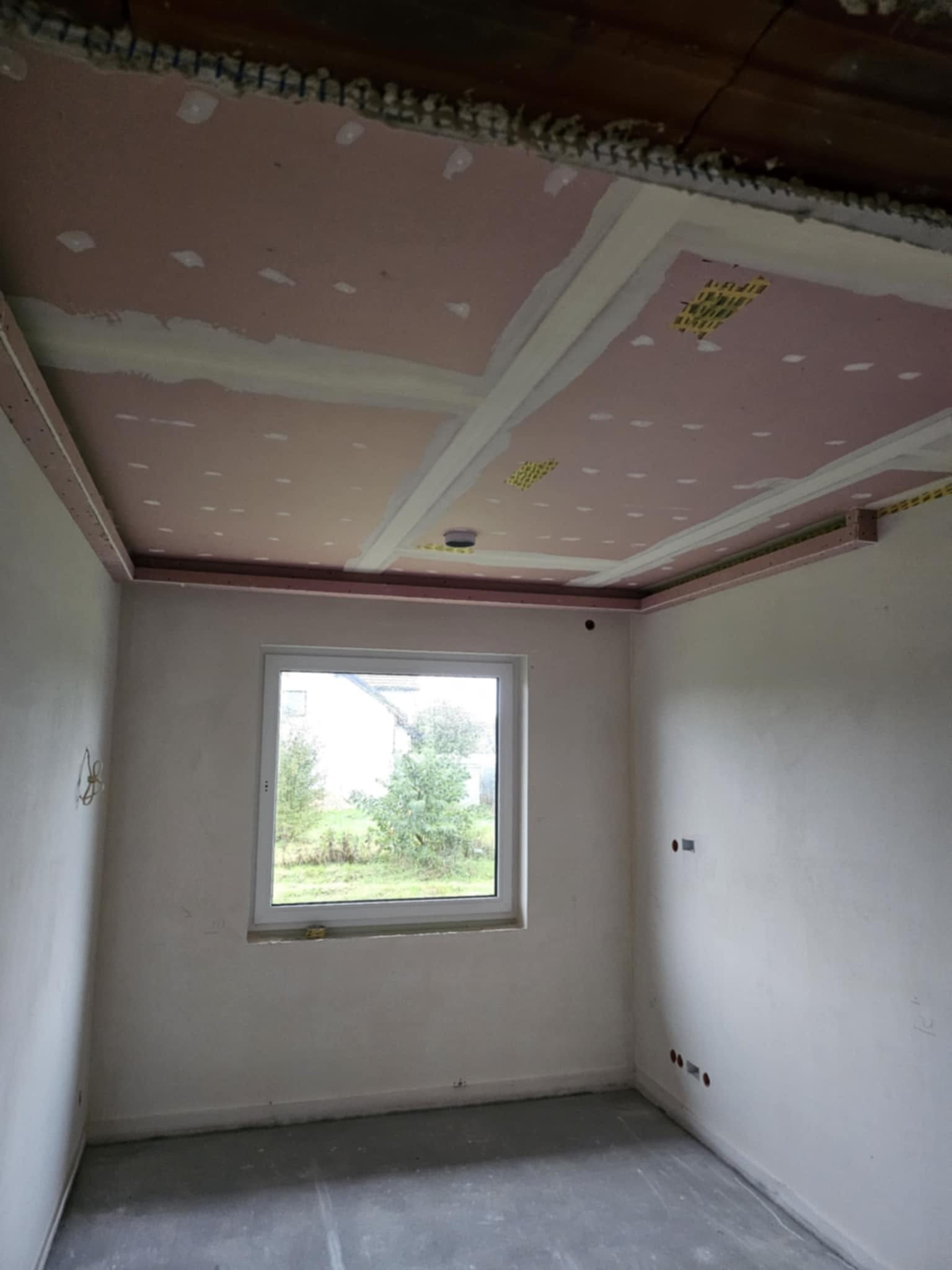 Pokój w trakcie prac wykończeniowych: widoczne ściany po szpachlowaniu, zamontowana konstrukcja sufitu podwieszanego z płyt kartonowo-gipsowych.