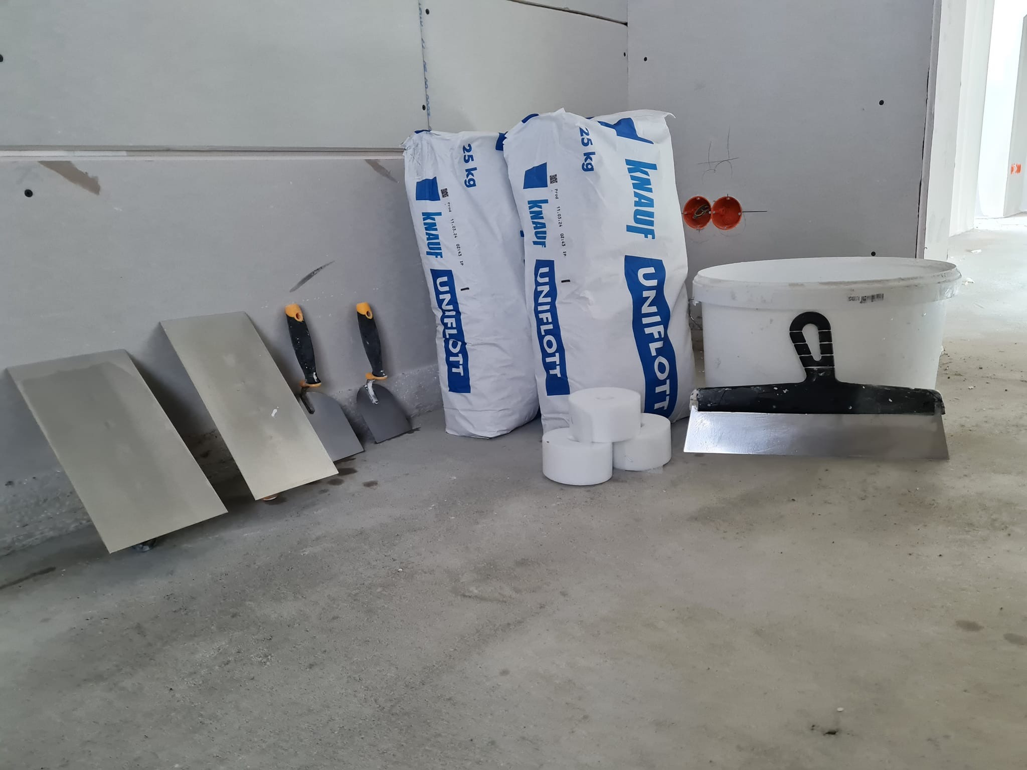 Przygotowane narzędzia i materiały budowlane: paka z masą szpachlową Knauf Uniflott, szpachelki i wiadro na tle ściany z płyt karton-gips, przed rozpoczęciem prac wykończeniowych.