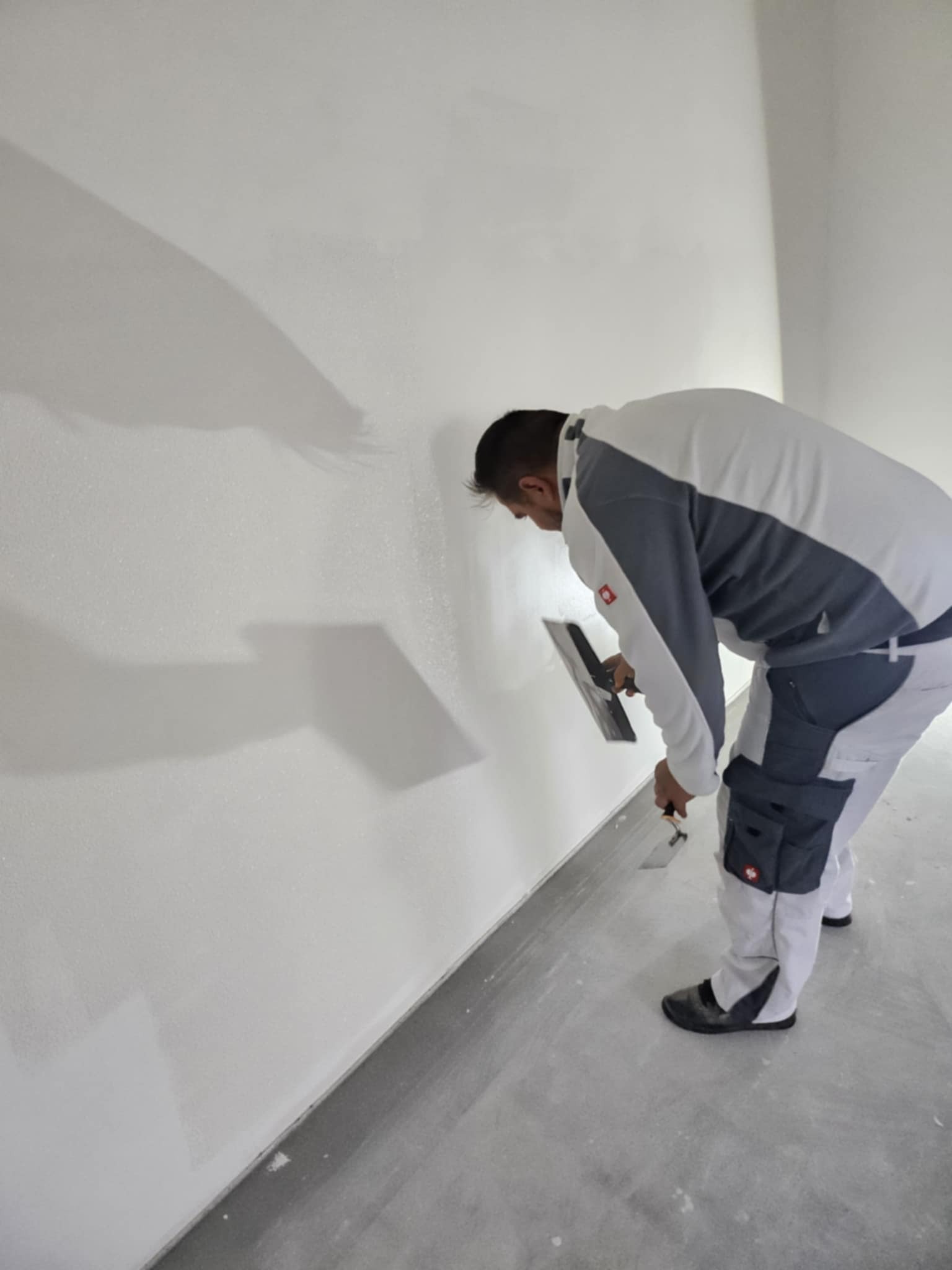 Pracownik w trakcie nakładania gładzi na ścianę w jasnym pomieszczeniu. Widoczne narzędzia: szpachelka i paca. Ściana częściowo pokryta gładzią, szary betonowy podkład.