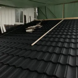 Czarny dach z blachodachówki modułowej w trakcie montażu, widoczne elementy konstrukcyjne i resztki materiałów, budynek z białym sidingiem w tle.