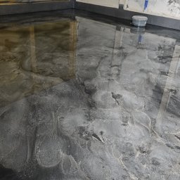 PERRY-BUD Patryk Ślusarski - Błyszcząca, szara posadzka żywiczna o marmurkowym wzorze w trakcie realizacji, widoczne ślady po narzędziach i wiaderko z farbą przy ścianie z niedokończonymi fragmentami gładzi.