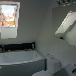 PERRY-BUD Patryk Ślusarski - Jasna łazienka na poddaszu z białą wanną, toaletą i umywalką, okno dachowe z widokiem na dach i ogród, ściany wyłożone białą, kwadratową mozaiką.