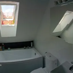 Jasna łazienka na poddaszu z białą wanną, toaletą i umywalką, okno dachowe z widokiem na dach i ogród, ściany wyłożone białą, kwadratową mozaiką.