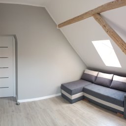 PERRY-BUD Patryk Ślusarski - Pokój na poddaszu z szarą ścianą, drewnianą konstrukcją stropu, jasną podłogą i narożną sofą w odcieniach szarości i bieli.