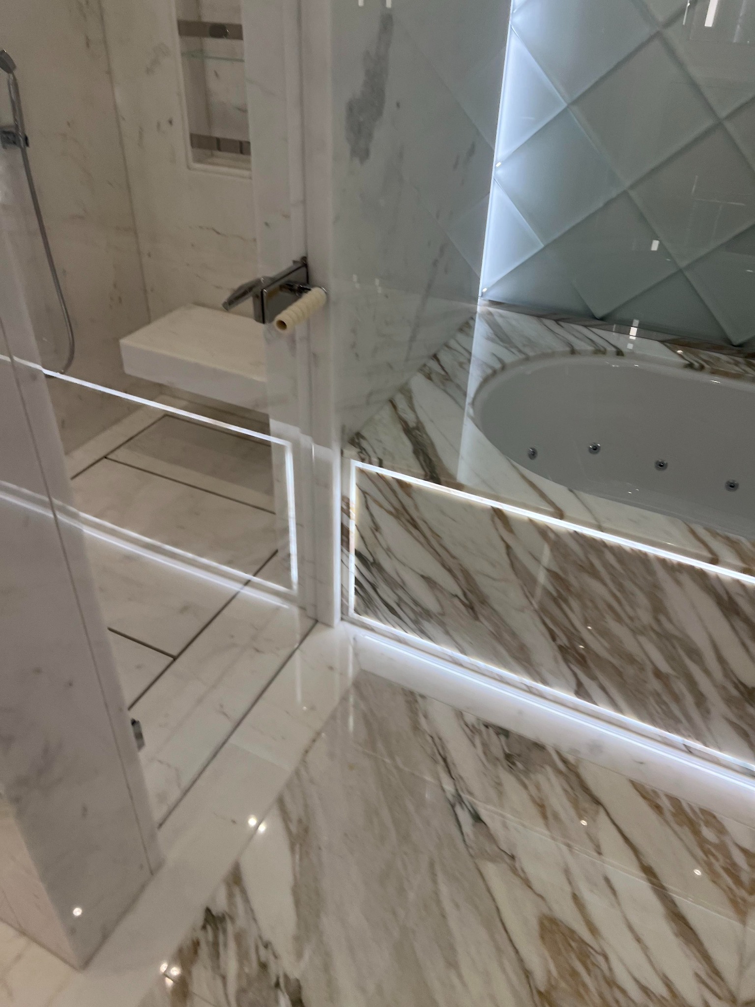 Nowoczesna łazienka z marmurową podłogą, ścianami i kabiną prysznicową ze szklanymi drzwiami z podświetleniem LED, widoczna wanna z hydromasażem i białe płytki na ścianie