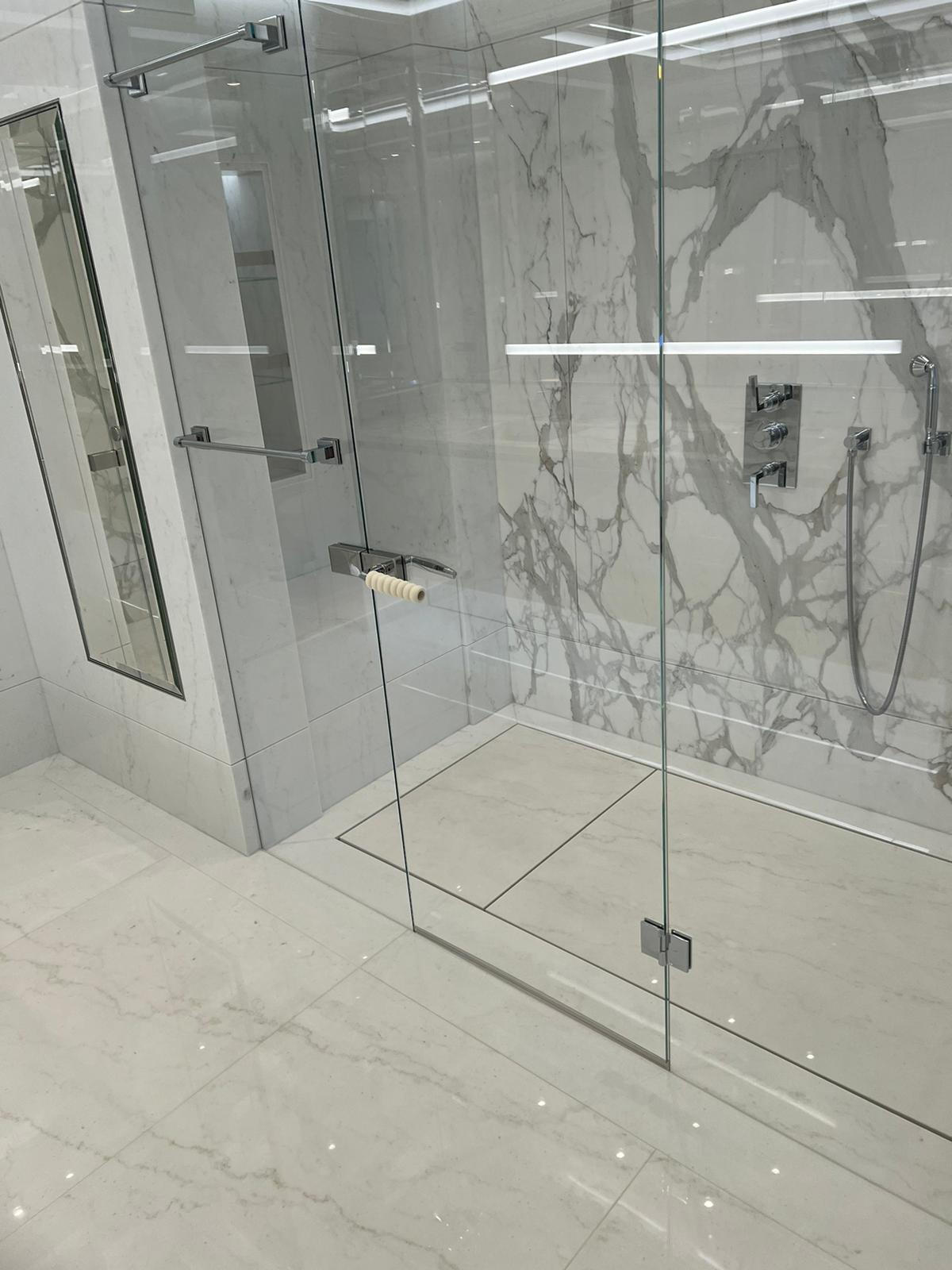 Nowoczesna kabina prysznicowa z białymi marmurowymi płytkami, chromowaną armaturą i szklanymi drzwiami z uchwytem z jasnego drewna.