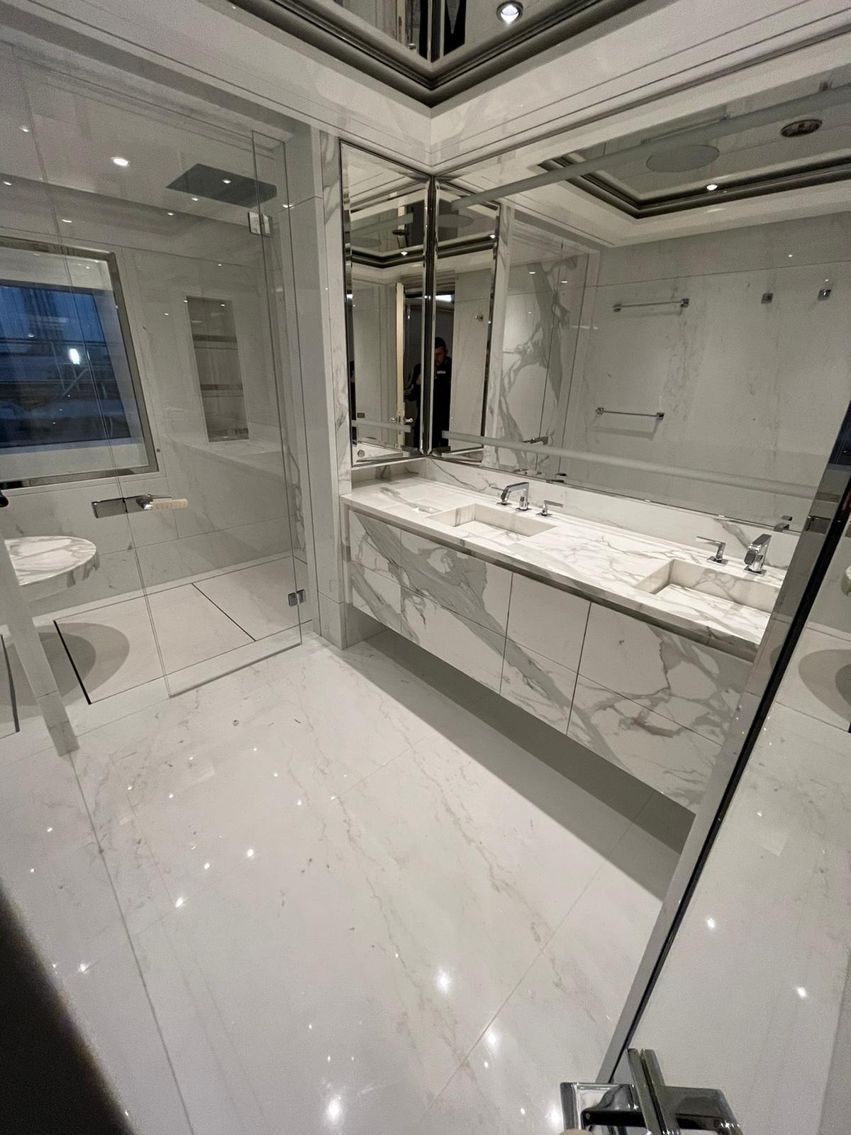 Elegancka, biała łazienka z marmurowym wzorem na podłodze, ścianach i szafce pod umywalką, kabina prysznicowa ze szklanymi drzwiami, duże lustra z chromowanymi ramami.