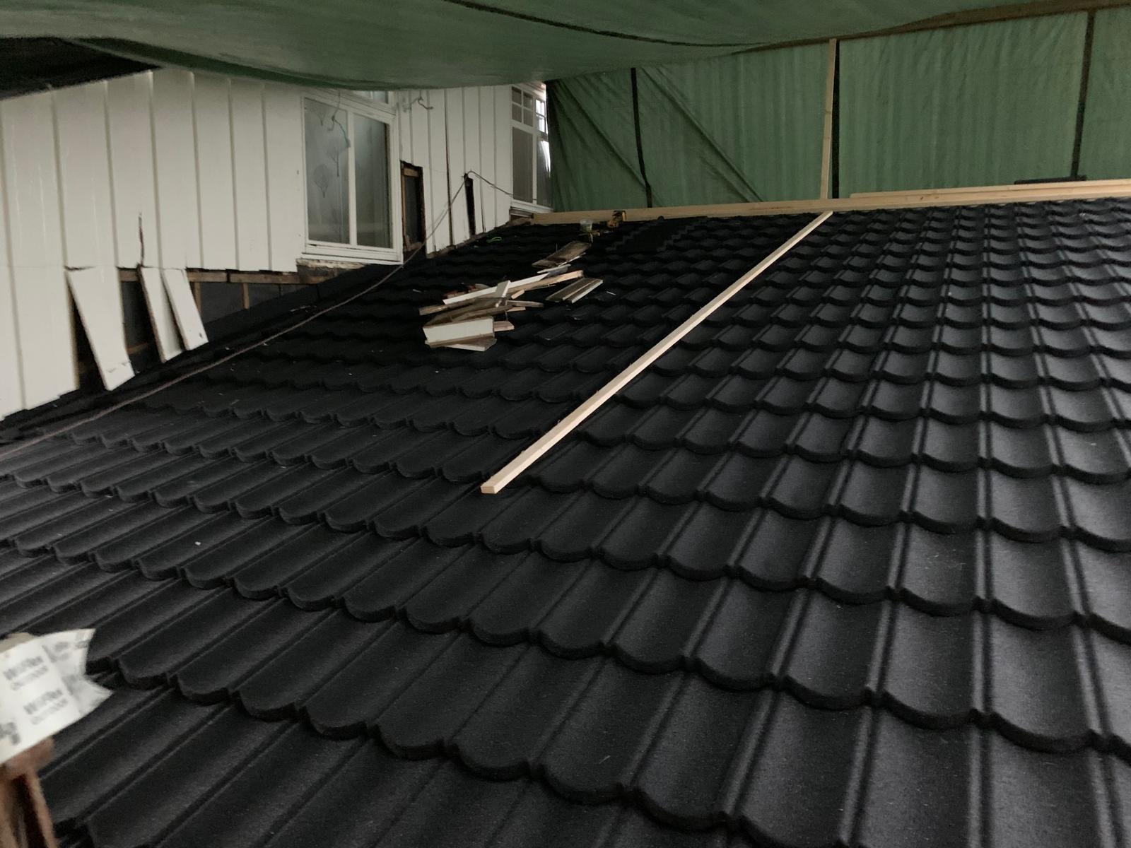 Czarny dach z blachodachówki modułowej w trakcie montażu, widoczne elementy konstrukcyjne i resztki materiałów, budynek z białym sidingiem w tle.