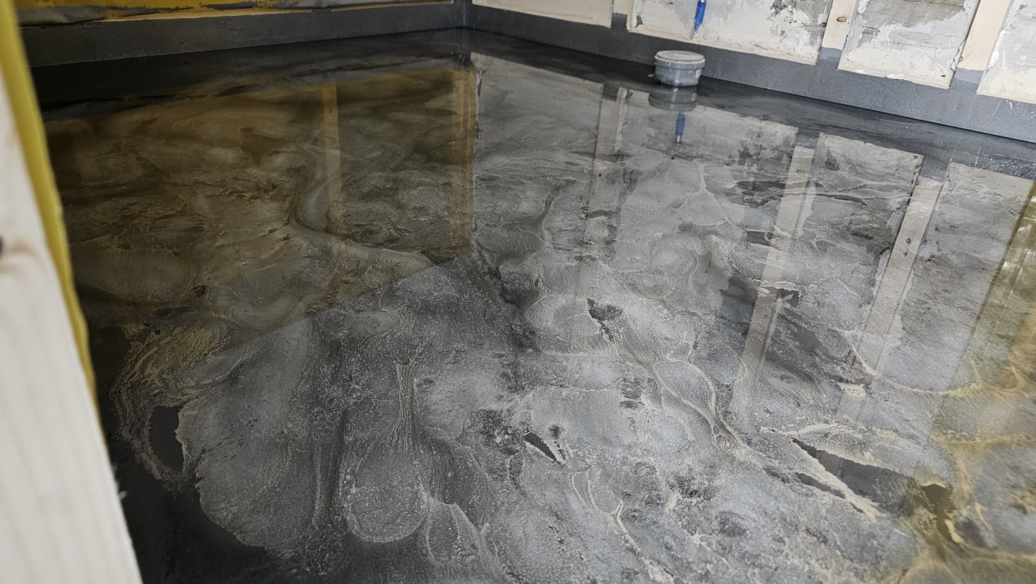 Błyszcząca, szara posadzka żywiczna o marmurkowym wzorze w trakcie realizacji, widoczne ślady po narzędziach i wiaderko z farbą przy ścianie z niedokończonymi fragmentami gładzi.