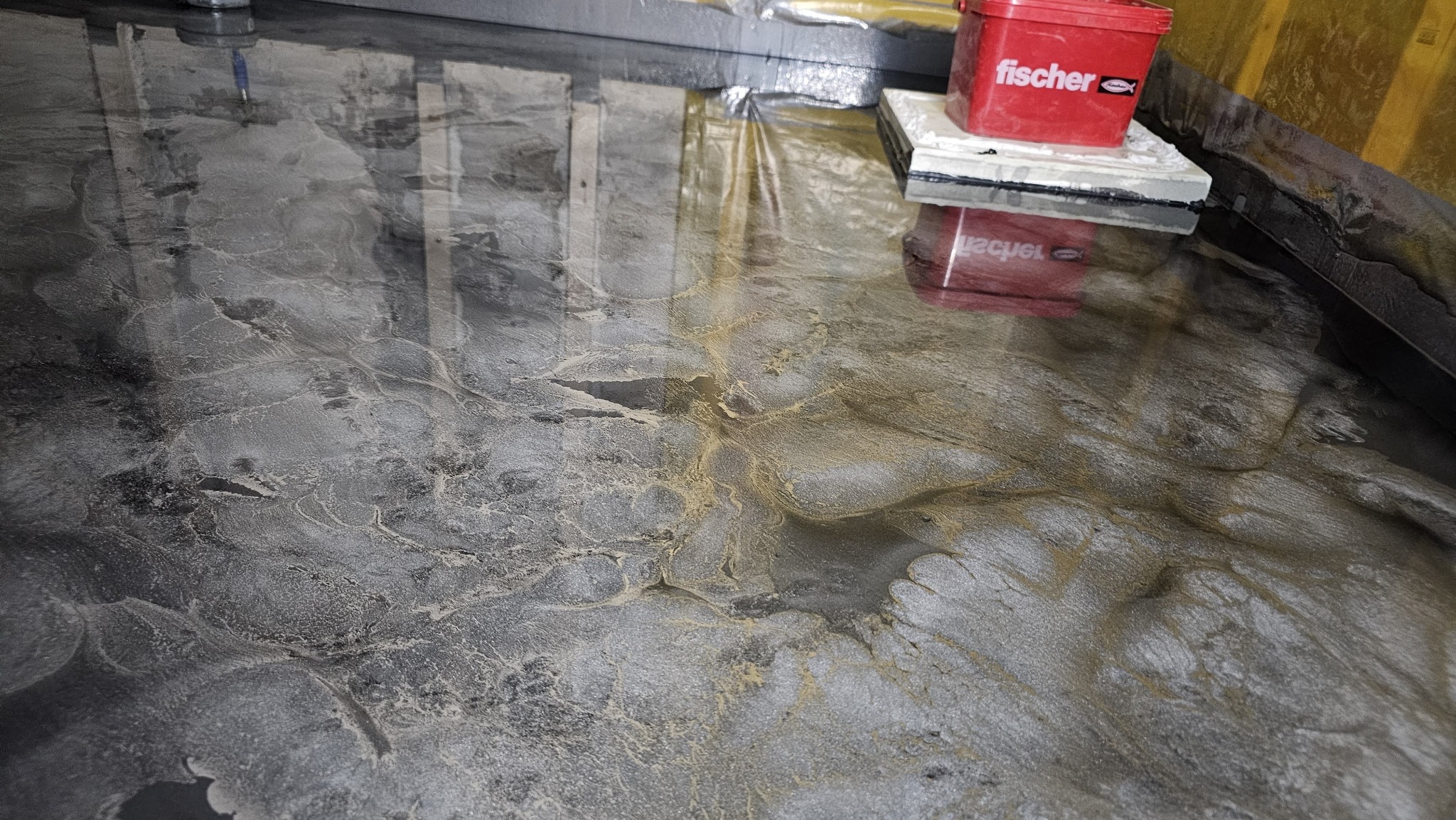 Błyszcząca, szaro-złota posadzka żywiczna z efektem marmuru, odbijająca światło i narzędzia pracy, z widocznym wiaderkiem firmy Fischer w rogu pomieszczenia.