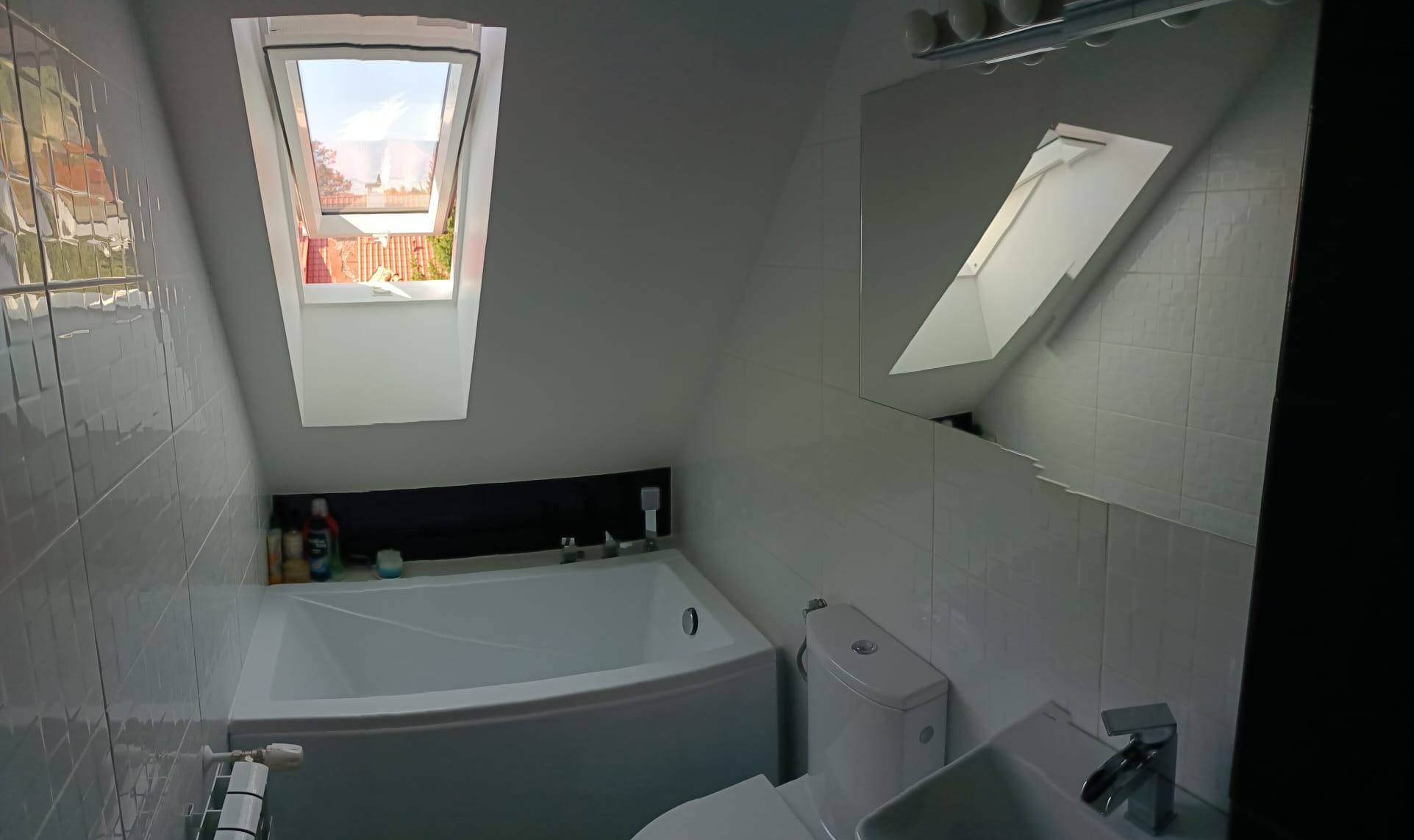 Jasna łazienka na poddaszu z białą wanną, toaletą i umywalką, okno dachowe z widokiem na dach i ogród, ściany wyłożone białą, kwadratową mozaiką.