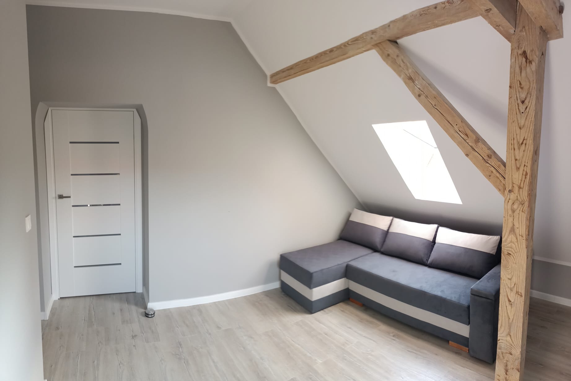Pokój na poddaszu z szarą ścianą, drewnianą konstrukcją stropu, jasną podłogą i narożną sofą w odcieniach szarości i bieli.