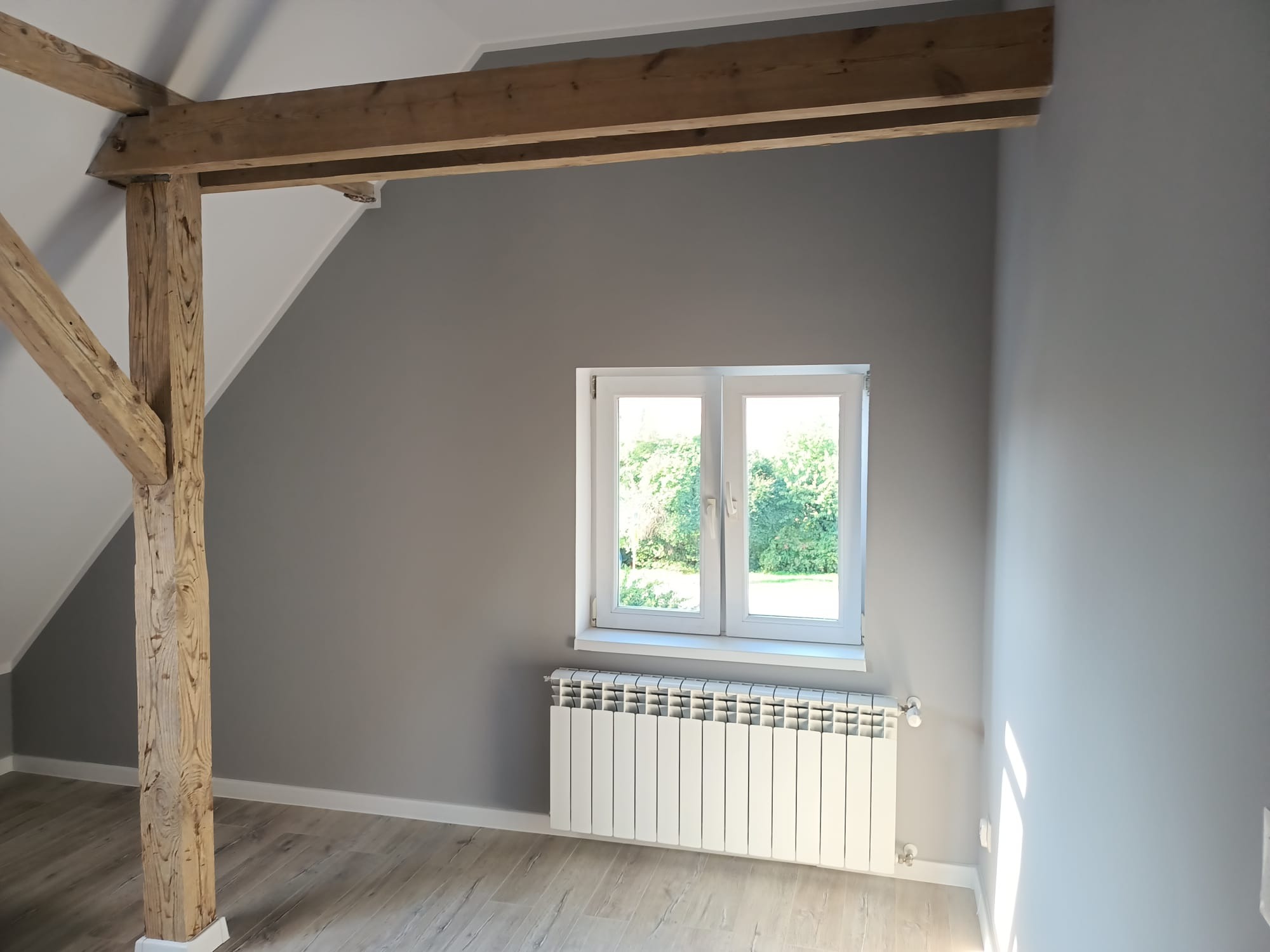 Szare wnętrze poddasza z drewnianą konstrukcją nośną, białym oknem i grzejnikiem, wykończone panelami podłogowymi.