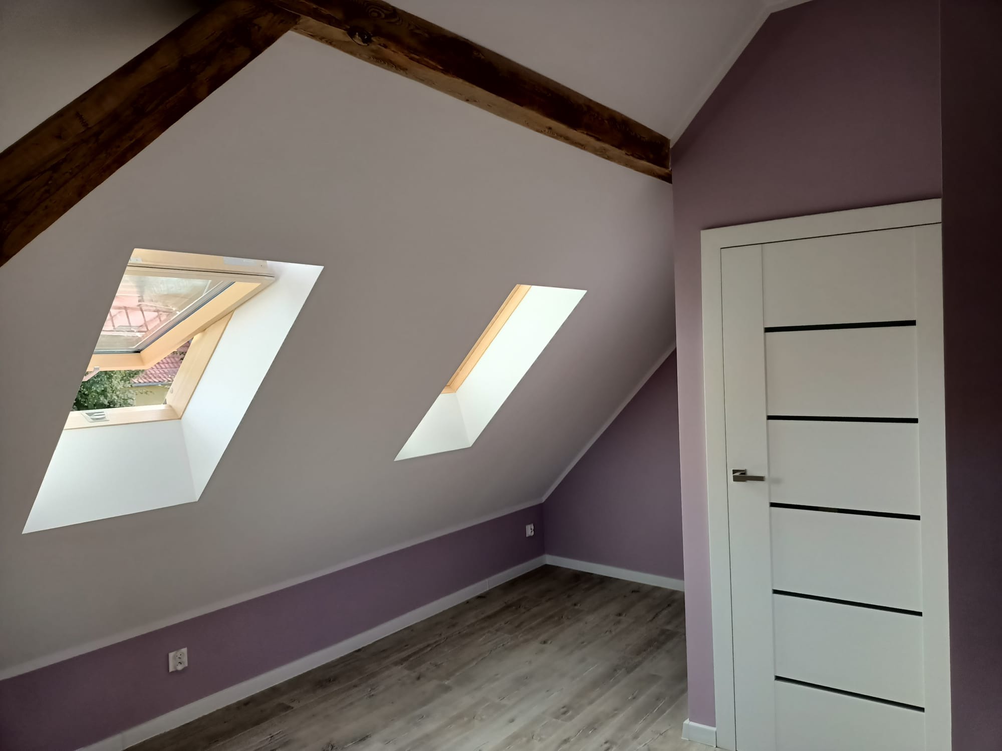 Poddasze po remoncie: Ściany pomalowane na fioletowo, białe okna dachowe z drewnianą ramą, drzwi wewnętrzne białe z czarnymi wstawkami, podłoga z paneli imitujących drewno, widoczne drewniane belki...