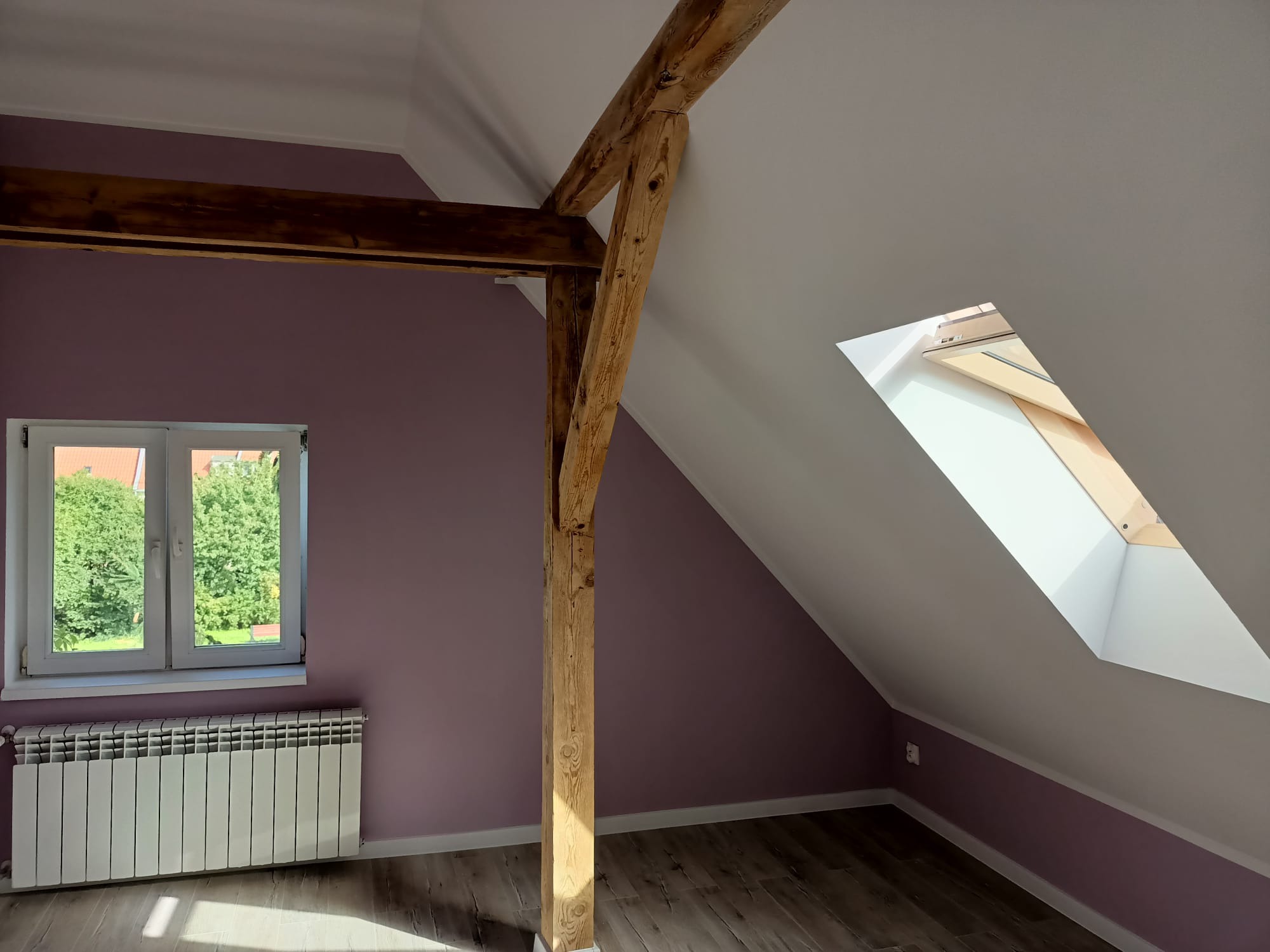 Poddasze po remoncie: fioletowe ściany, drewniana belka konstrukcyjna, okno dachowe i grzejnik pod oknem.