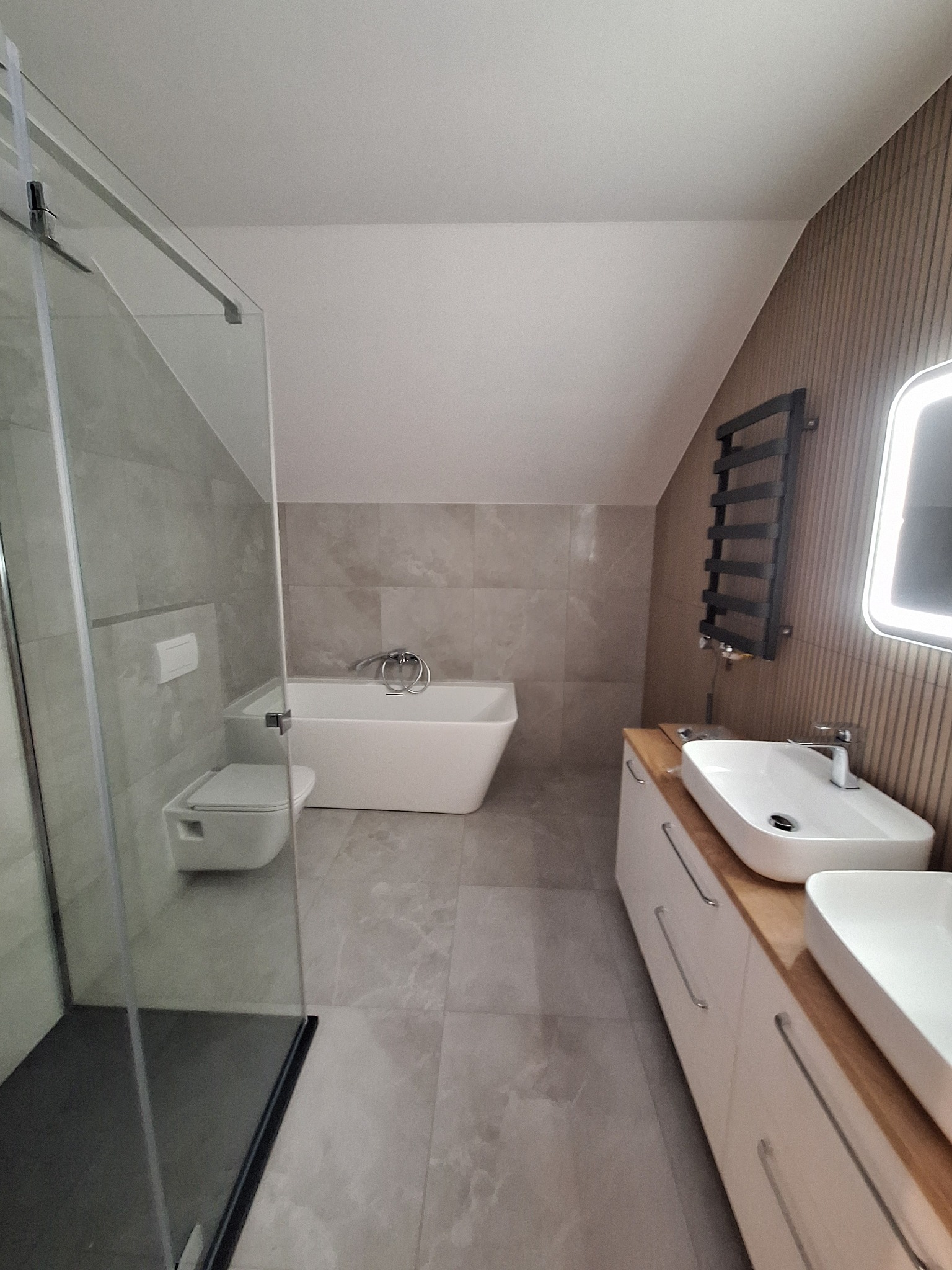 Nowoczesna łazienka na poddaszu z kabiną prysznicową typu walk-in, wolnostojącą wanną, podwójną umywalką nablatową na szafce z drewnianym blatem oraz grzejnikiem drabinkowym.