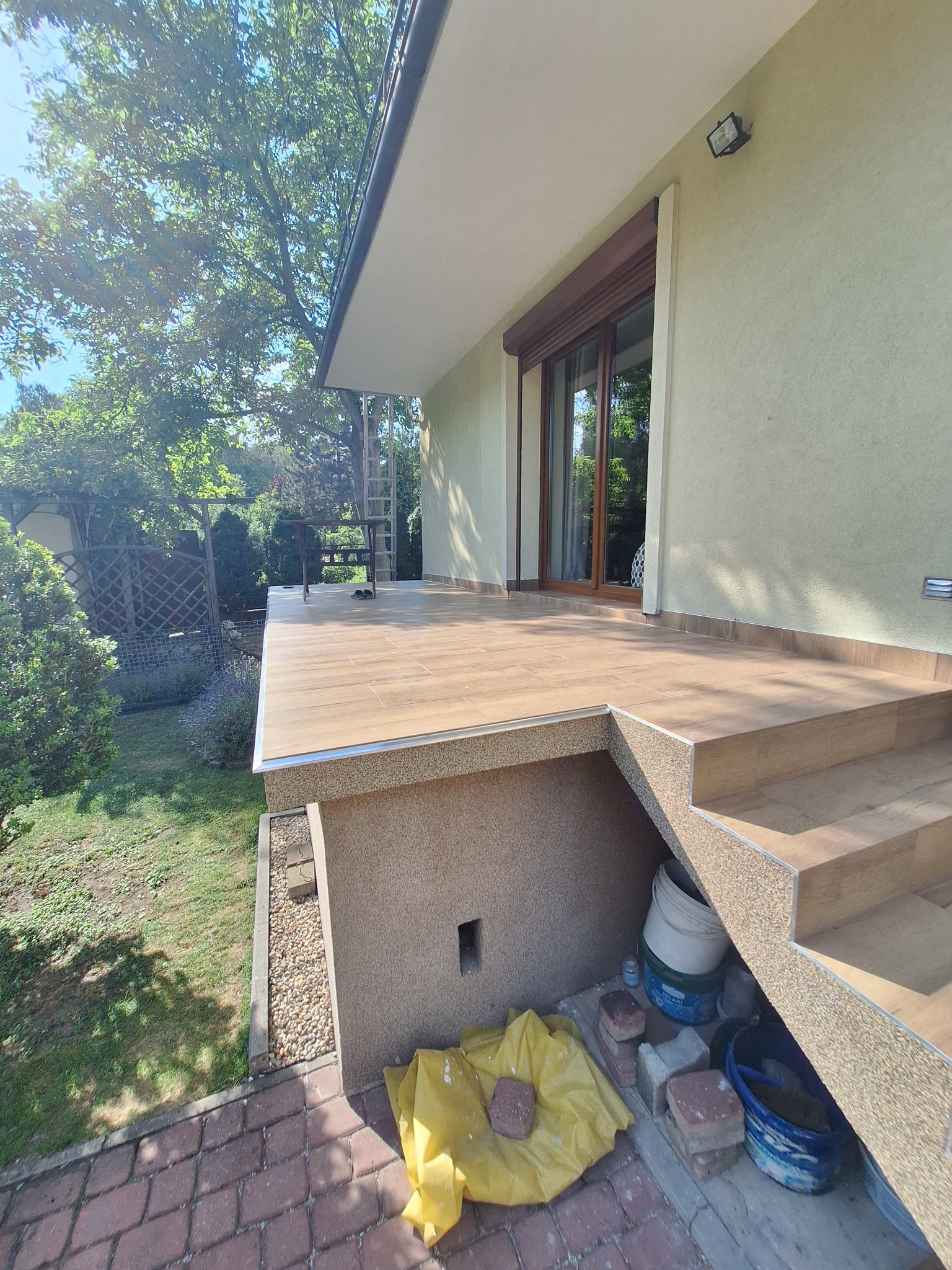 Wykończony taras z płytkami imitującymi drewno, schody z tego samego materiału, widoczne narzędzia i materiały budowlane pod schodami, otoczenie z zielenią i kostką brukową.