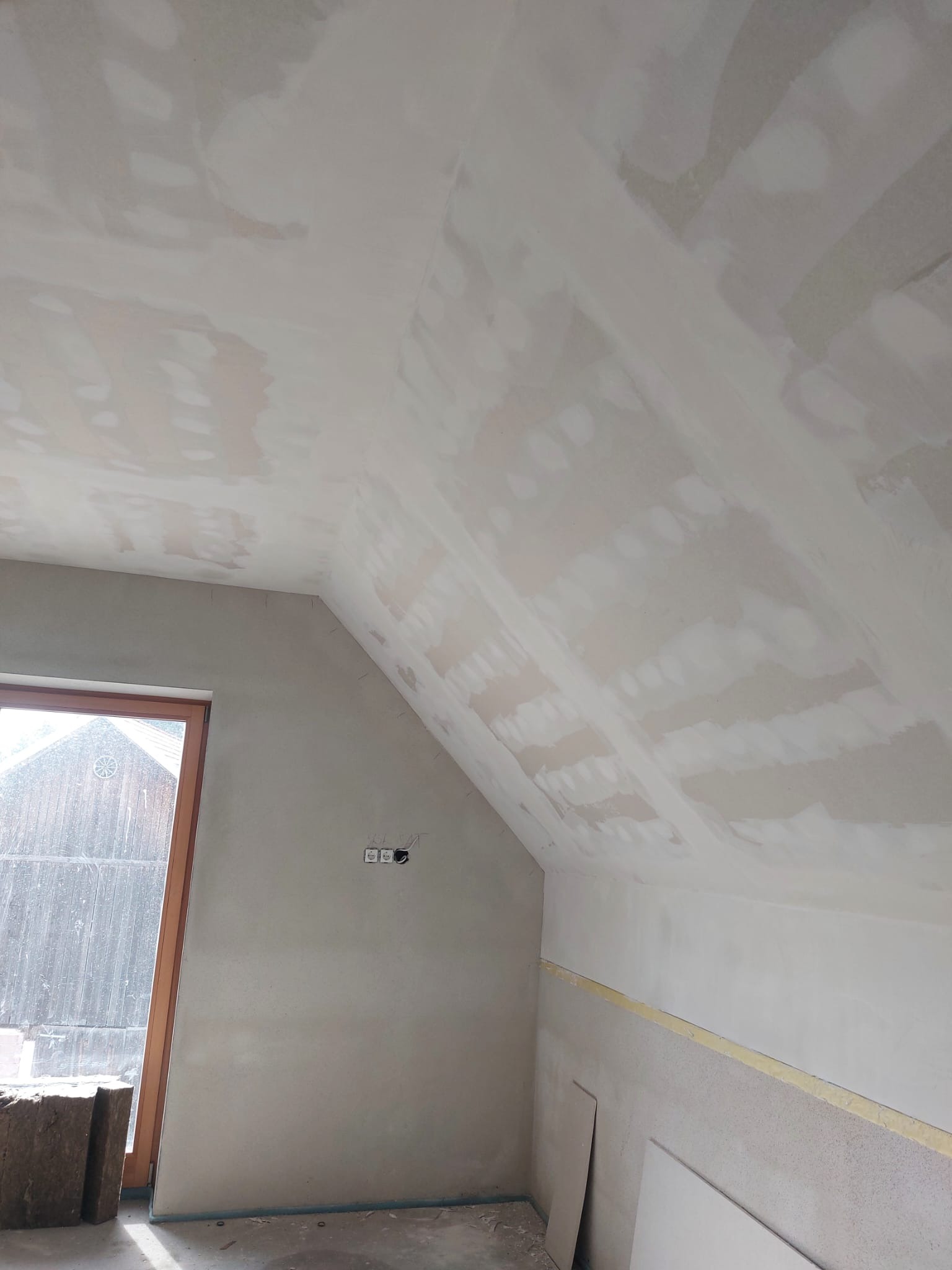 Poddasze w trakcie prac wykończeniowych: ściany i skośny sufit pokryte warstwą szpachli, widoczne puszki elektryczne, okno z drewnianą ramą i płyty gipsowe przy ścianie.