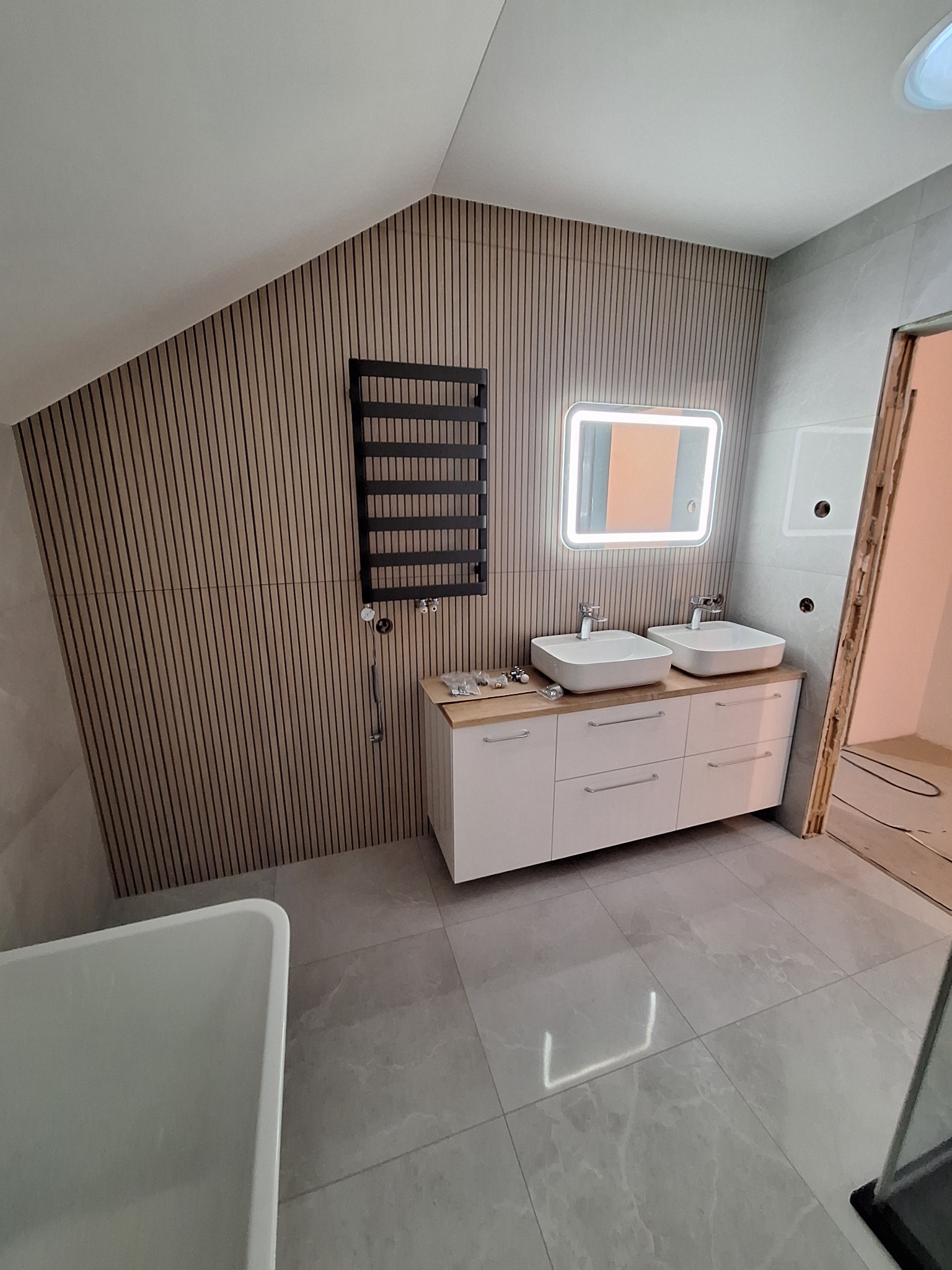 Nowoczesna łazienka z drewnianymi panelami ściennymi, podwójną umywalką na szafce z jasnego drewna i podświetlanym lustrem, grzejnik drabinkowy na ścianie, szare płytki podłogowe.