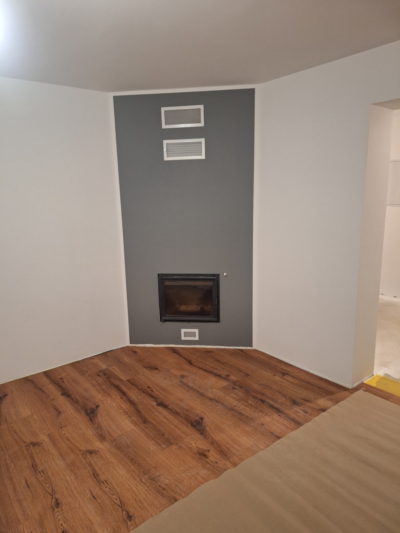 Nowoczesny salon po wykończeniu: drewniana podłoga, szara ściana z kominkiem i białymi akcentami. Widok na sąsiednie pomieszczenie.