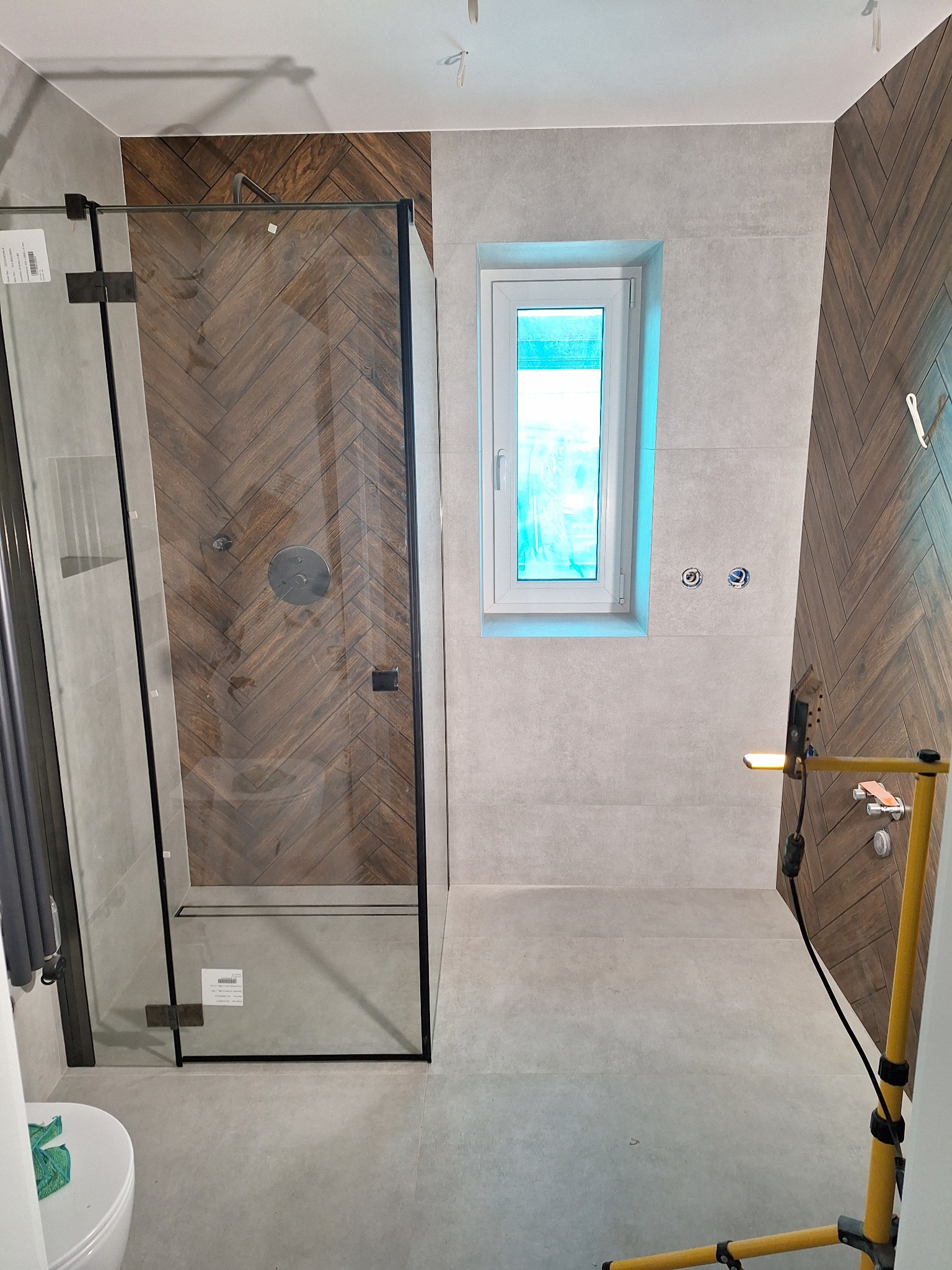 Wykończona łazienka z kabiną prysznicową typu walk-in, wyłożona szarą płytką i drewnopodobną w jodełkę. Widoczne przyłącza wodne i fragment toalety. Oświetlenie budowlane.