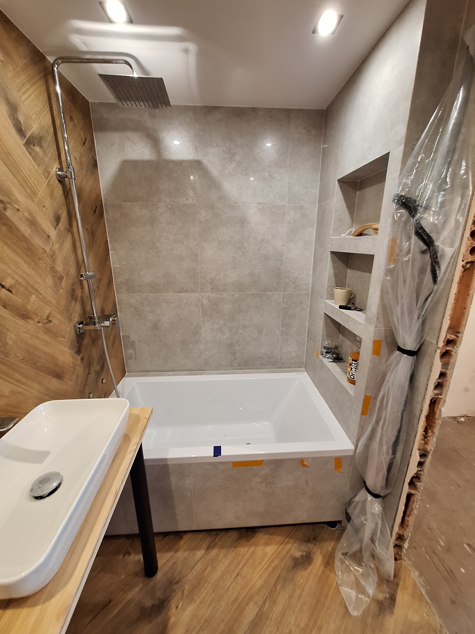 Nowoczesna łazienka z drewnianą ścianą, szarą armaturą i wnękami w ścianie. Umywalka na drewnianym blacie. Część ściany zabezpieczona folią malarską.