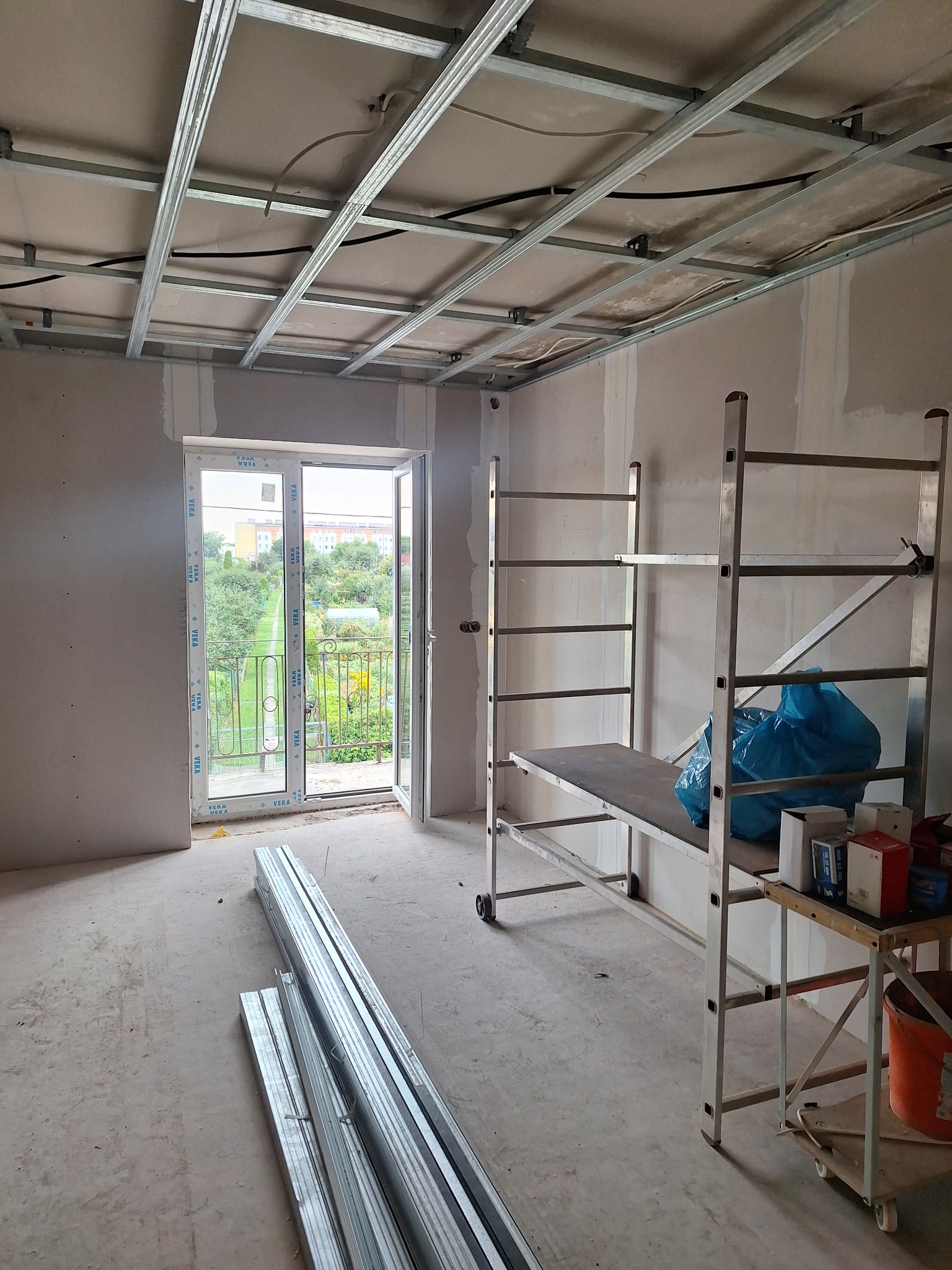 Wnętrze w trakcie prac wykończeniowych: metalowa konstrukcja sufitu podwieszanego, szpachlowane ściany, rusztowanie i profile. Widok na balkon z zielenią w tle.