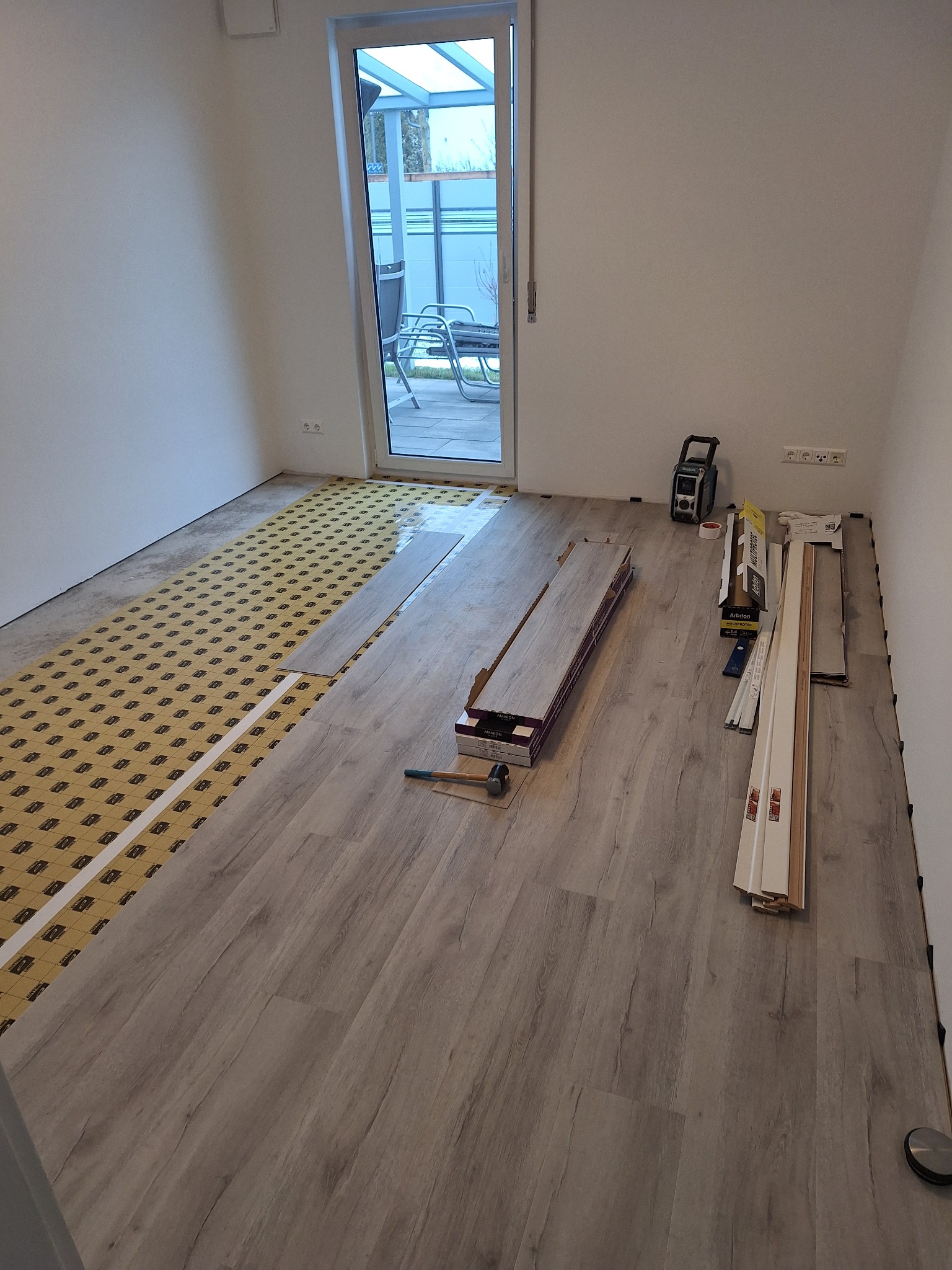 Układanie paneli podłogowych w jasnym pomieszczeniu. Częściowo ułożone panele, widoczna podkładka, narzędzia i listwy przypodłogowe. Prace w toku, widok na taras.