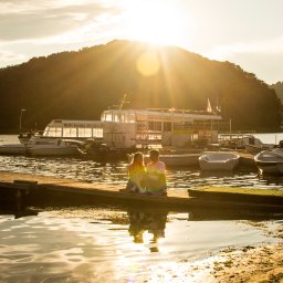 Czułe Kadry. Paulina Pluta - Fotografia - Para siedzi na pomoście nad jeziorem w promieniach zachodzącego słońca, w tle góra i łodzie. Romantyczna sceneria, ciepłe światło.