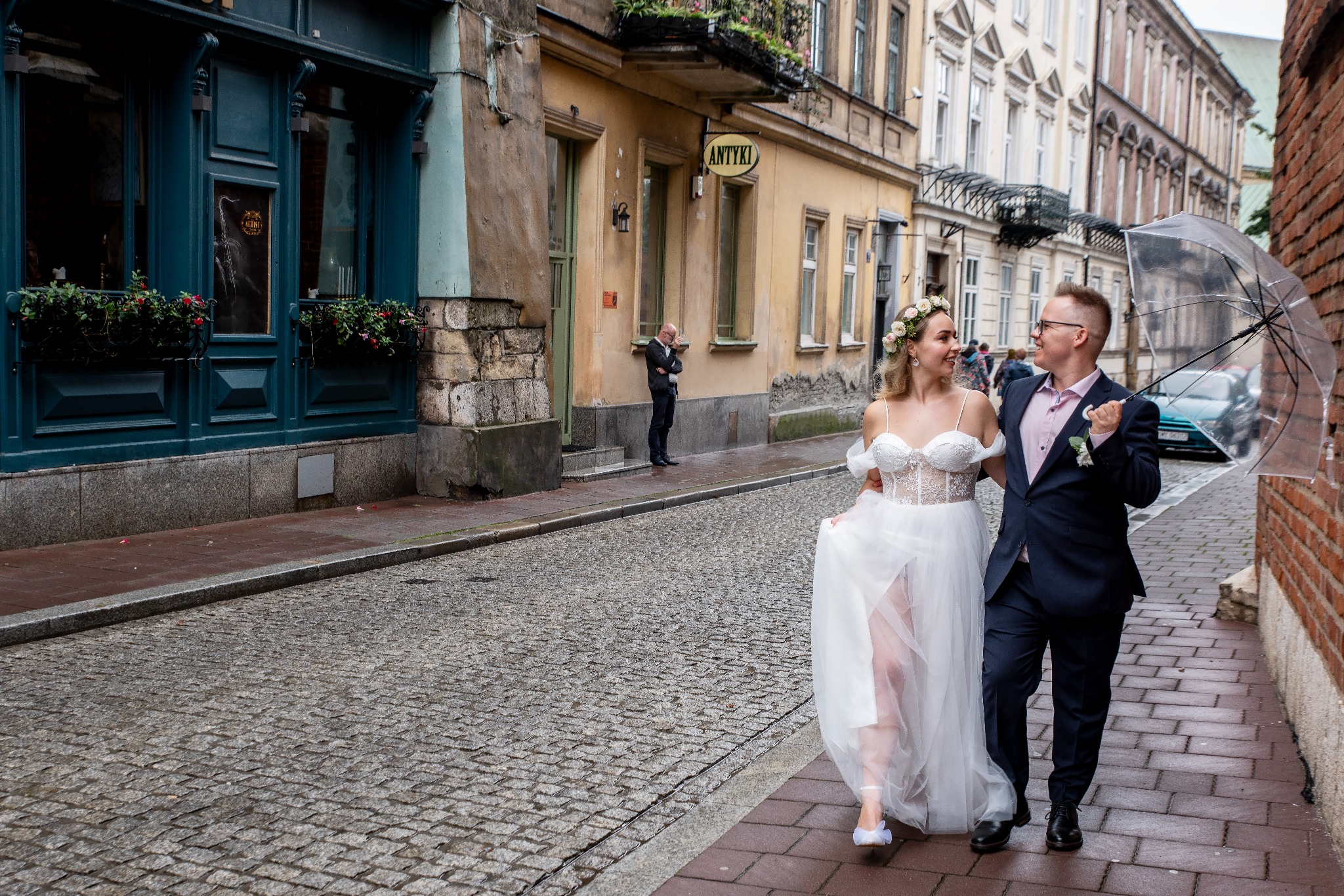 Para młoda spaceruje po brukowanej ulicy w Krakowie, trzymając parasol. W tle fotograf i budynki z napisem 'Antyki'.