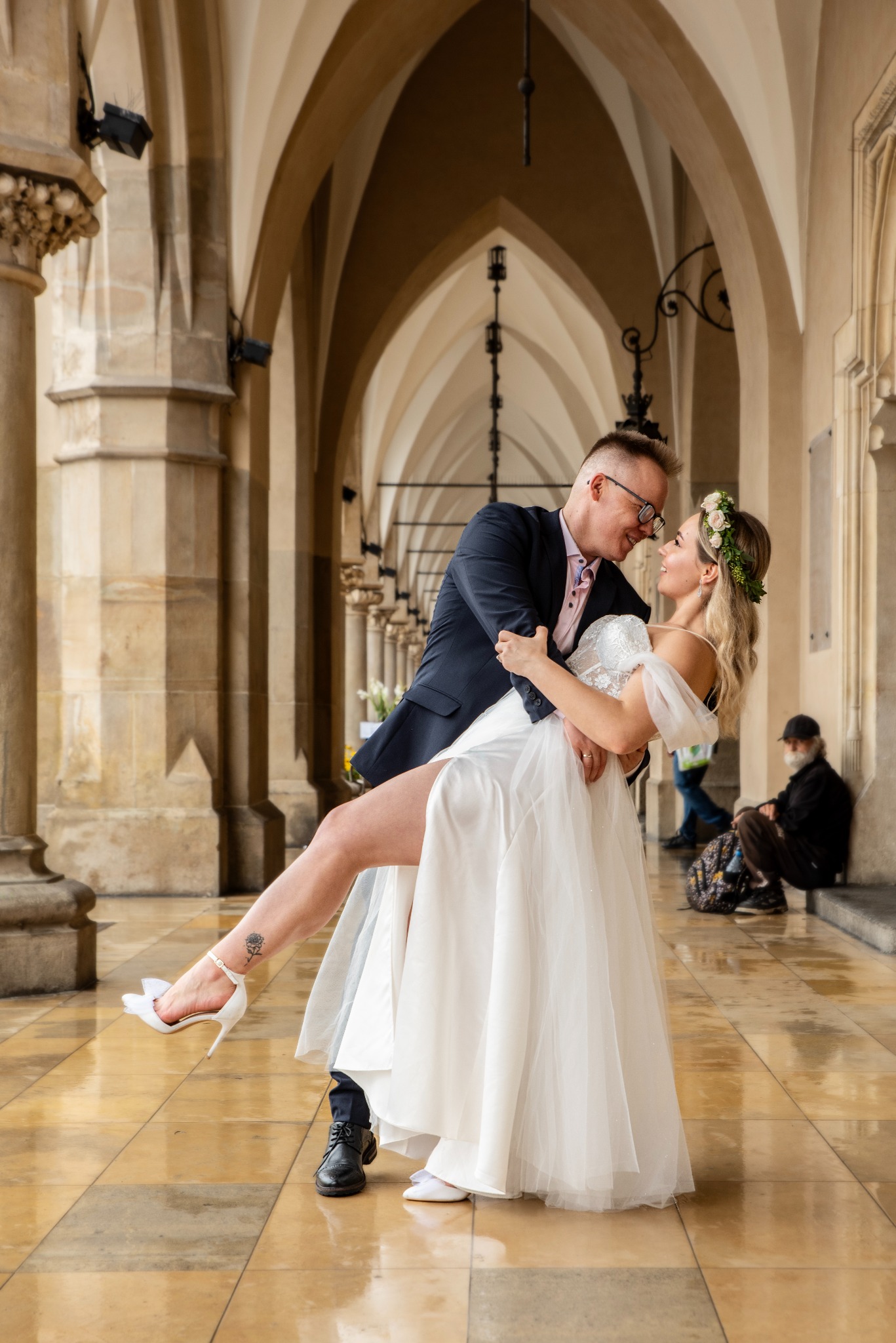 Para młoda w tańcu w architektonicznej scenerii. Panna młoda w sukni i wianku, pan młody w garniturze. Uśmiechnięci, zapatrzeni w siebie. W tle kolumnada.