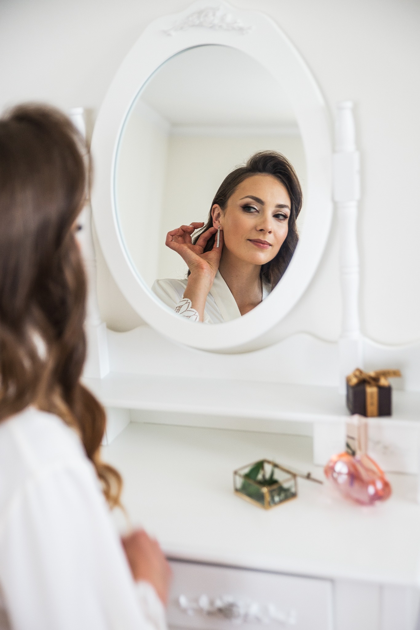 Panna młoda w białym szlafroku, przeglądająca się w owalnym lustrze toaletki, dopinająca kolczyk. Na blacie flakon perfum, pudełko i szkatułka.