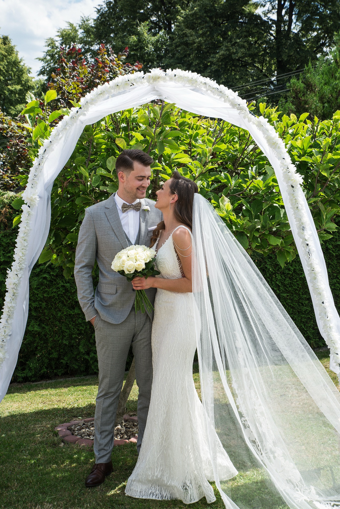 Para młoda w ogrodzie pod białym, kwiatowym łukiem. Panna młoda trzyma bukiet białych róż, oboje patrzą na siebie z uśmiechem. Ujęcie w słoneczny dzień.