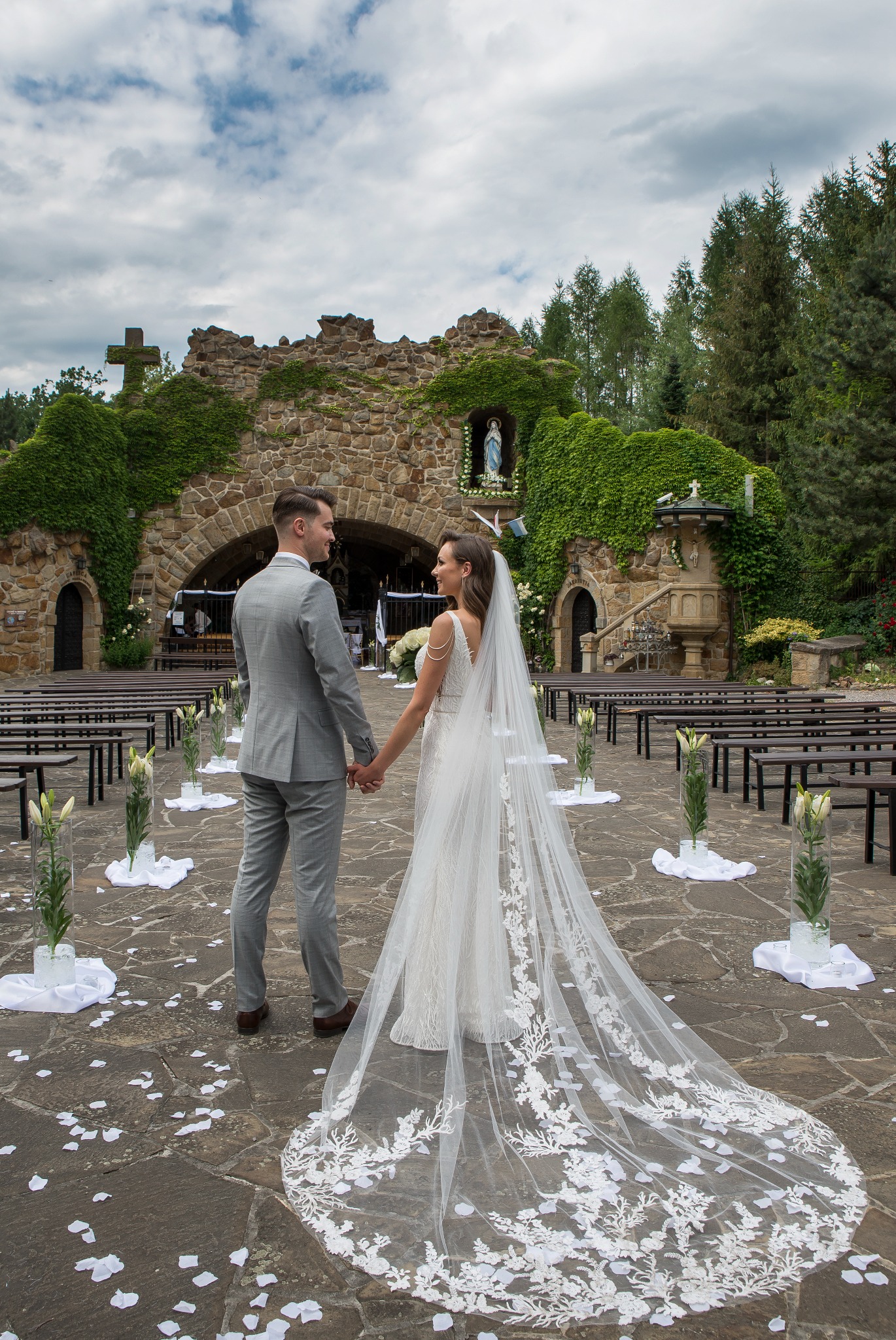 Para młoda, trzymając się za ręce, stoi tyłem na tle kamiennej kaplicy obrośniętej bluszczem. Długi welon panny młodej zdobiony jest koronką.