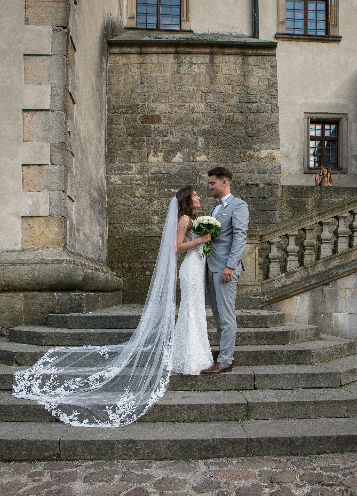 Para młoda w eleganckich strojach stoi na kamiennych schodach zamku. Długi, koronkowy welon panny młodej opada na stopnie. Romantyczny nastrój i architektura w tle.