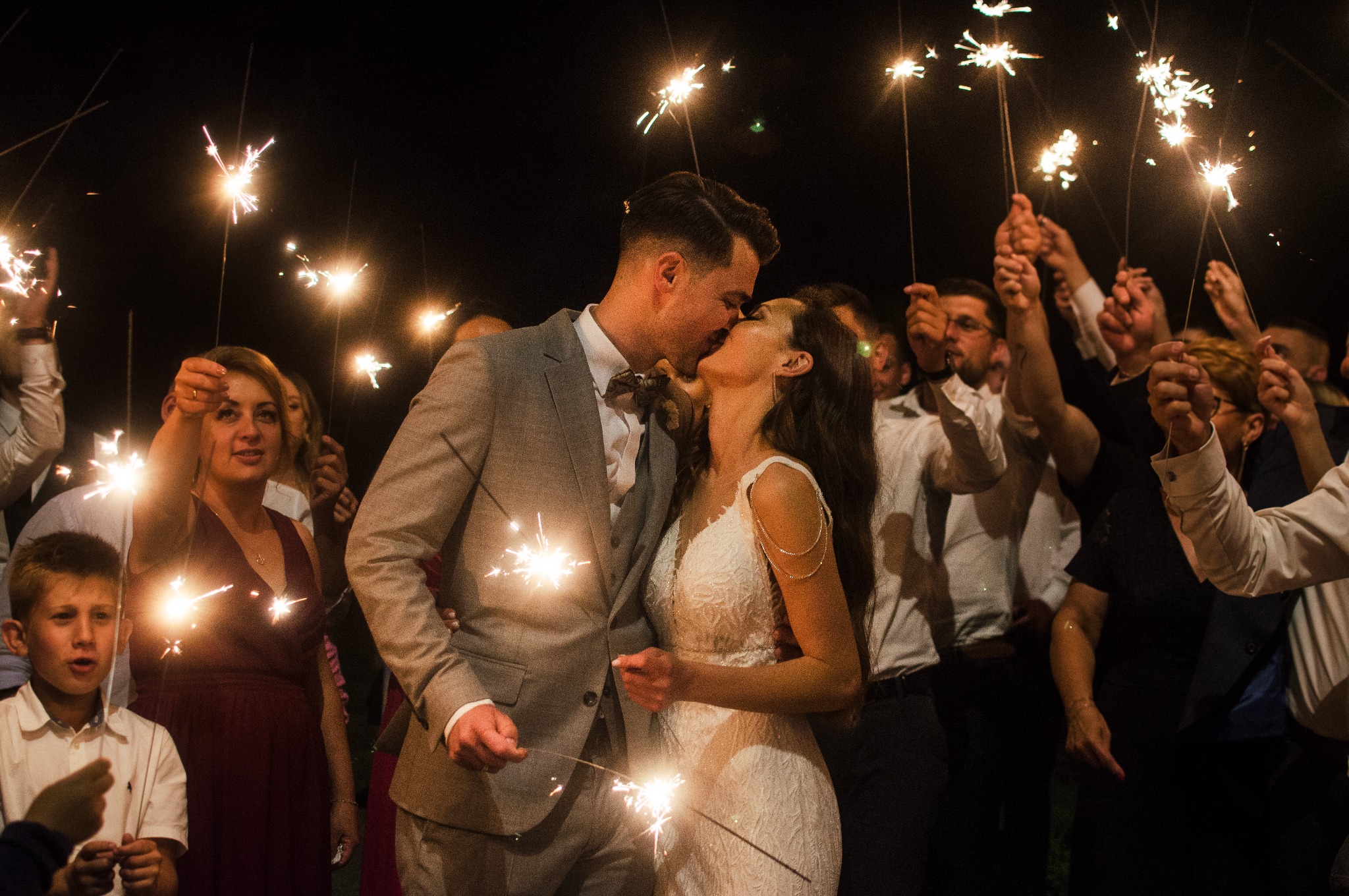 Pocałunek pary młodej otoczonej gośćmi z zimnymi ogniami w ciemności, subtelne oświetlenie, szary garnitur pana młodego i biała sukienka panny młodej.