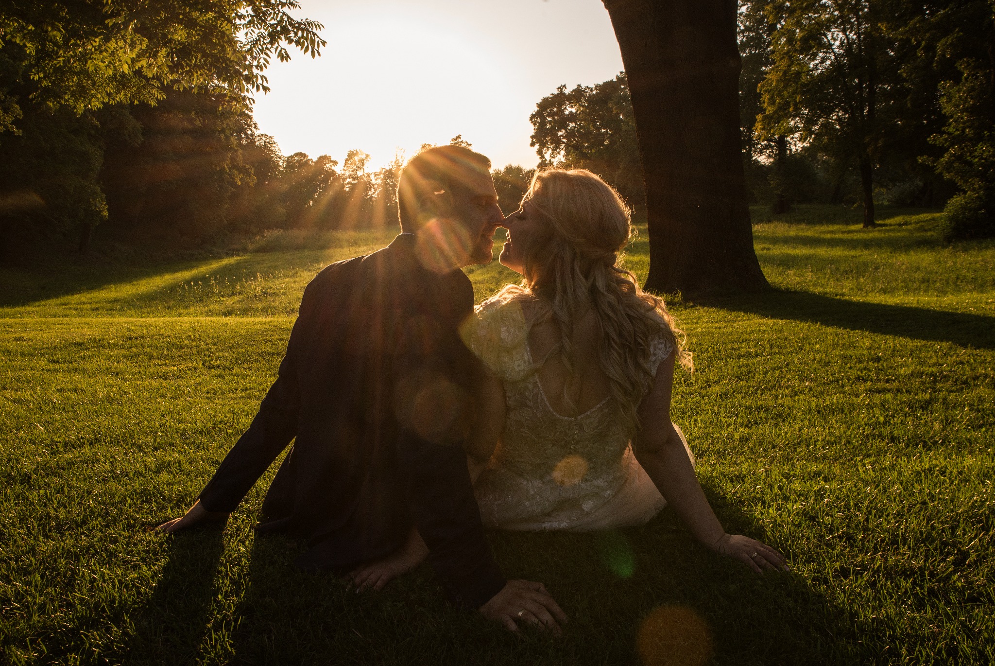 Para młoda siedzi na trawie w promieniach zachodzącego słońca. Sylwetki nowożeńców na tle drzew i zieleni, romantyczny nastrój, delikatne światło.