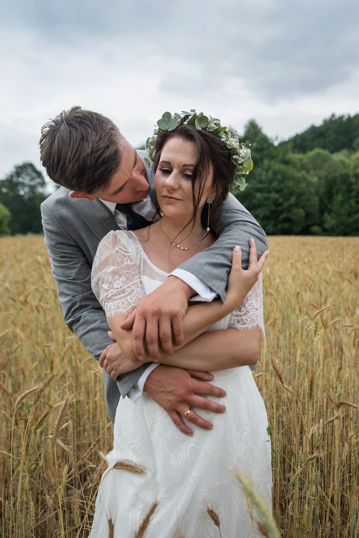 Młoda para w zbożu: Panna Młoda w wianku i koronkowej sukni, obejmowana przez Pana Młodego w szarym garniturze. Romantyczny kadr z zielonym tłem.