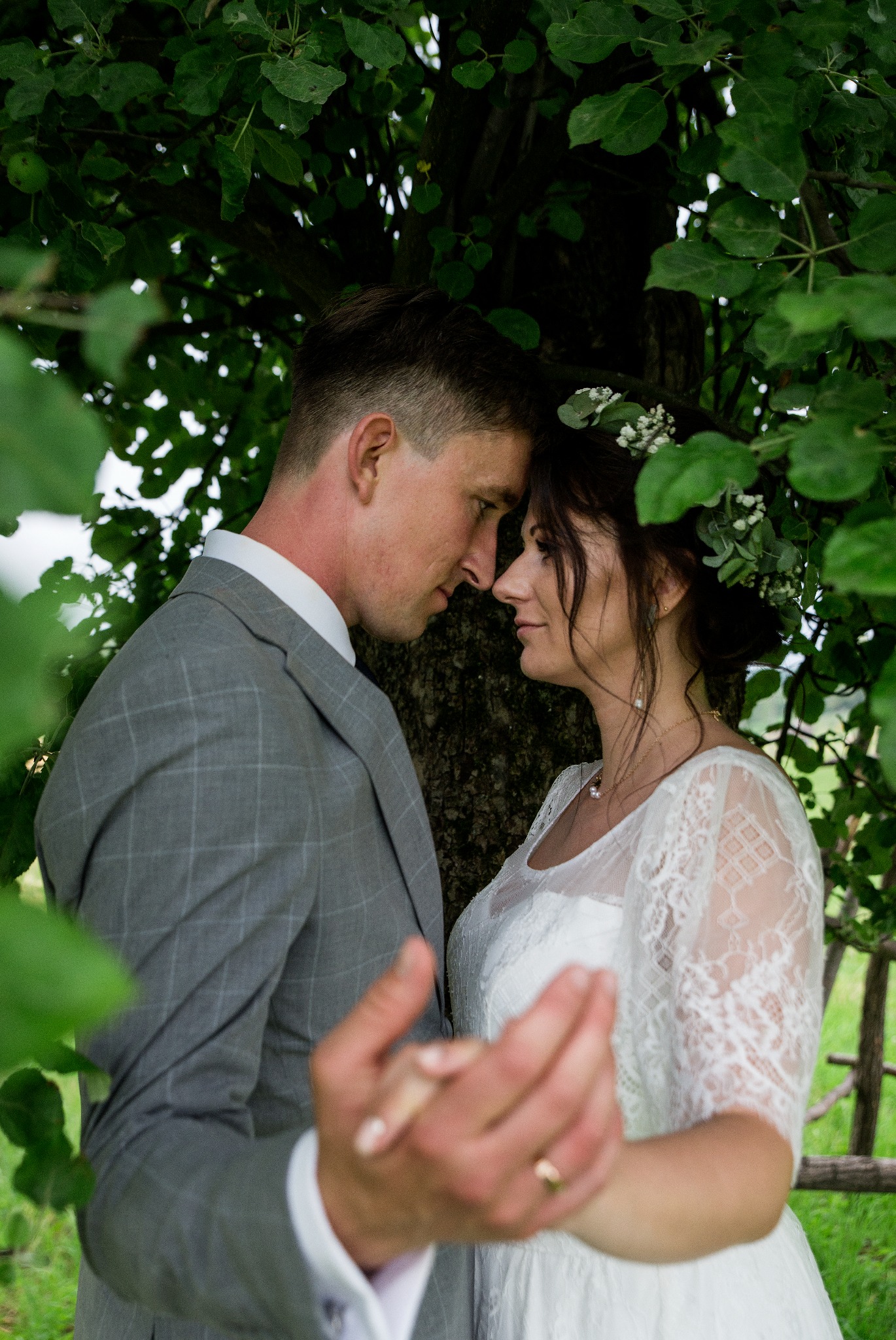 Para młoda w zbliżeniu, twarzami do siebie, pod drzewem. Panna młoda w wianku z białych kwiatów i koronkowej sukni. Pan młody w szarej marynarce.