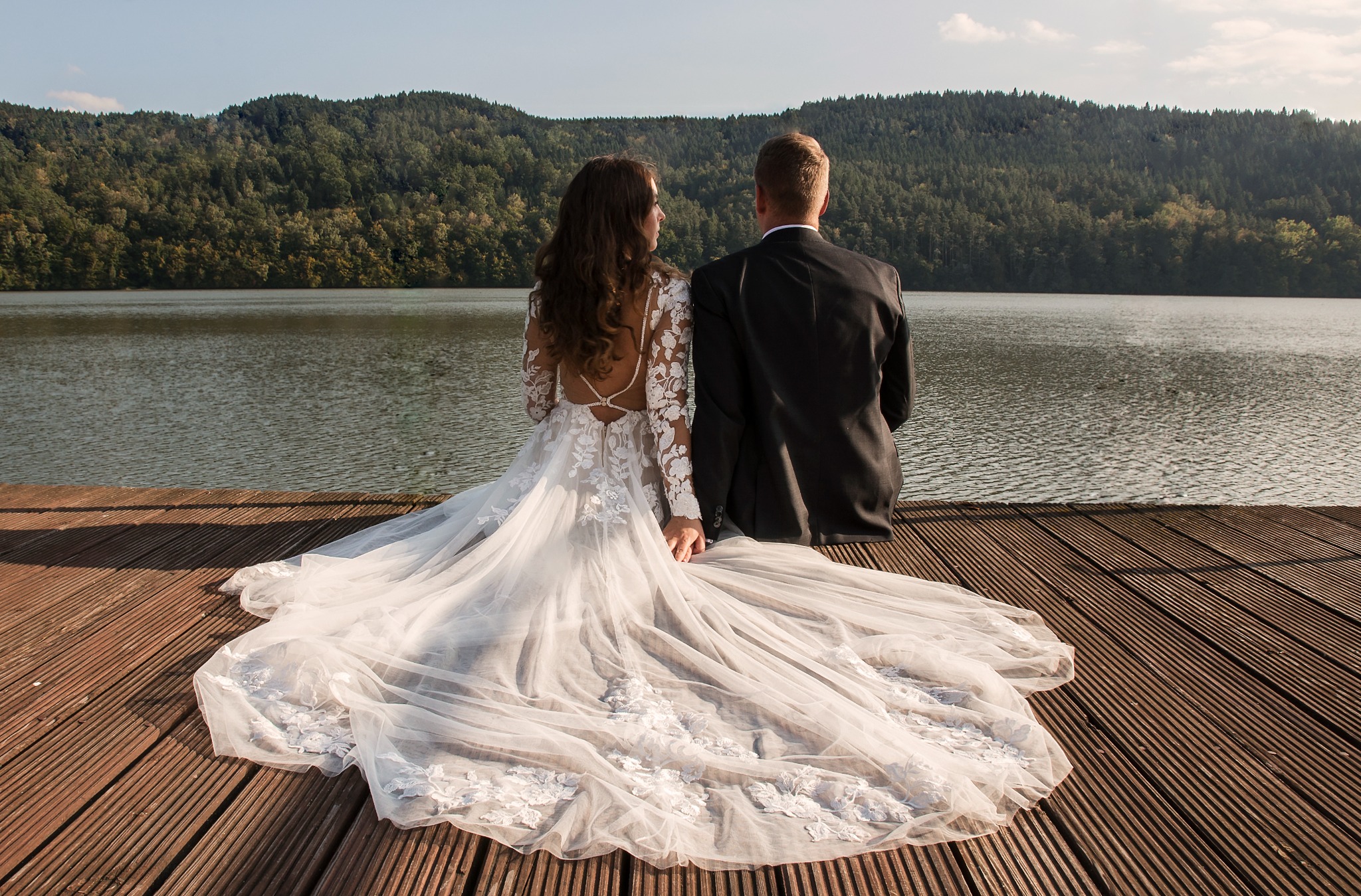 Para młoda siedzi na drewnianym molo, tyłem do kamery, z widokiem na jezioro i zalesione wzgórza w tle. Suknia panny młodej z koronkowymi aplikacjami.
