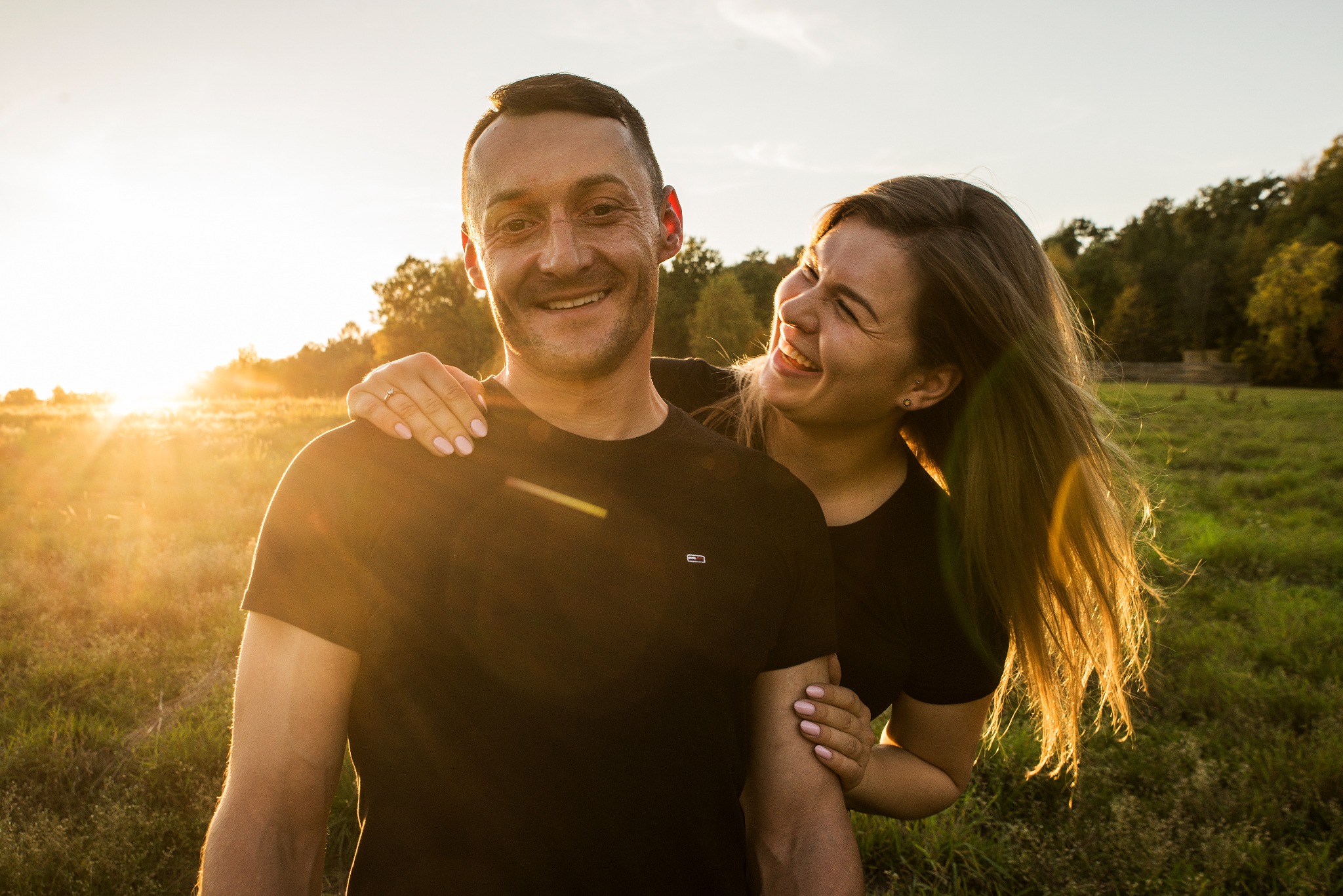 Zakochana para w Bochni, uśmiechnięta kobieta obejmuje mężczyznę na tle zachodzącego słońca i drzew. Ciepłe, naturalne światło podkreśla radość chwili.