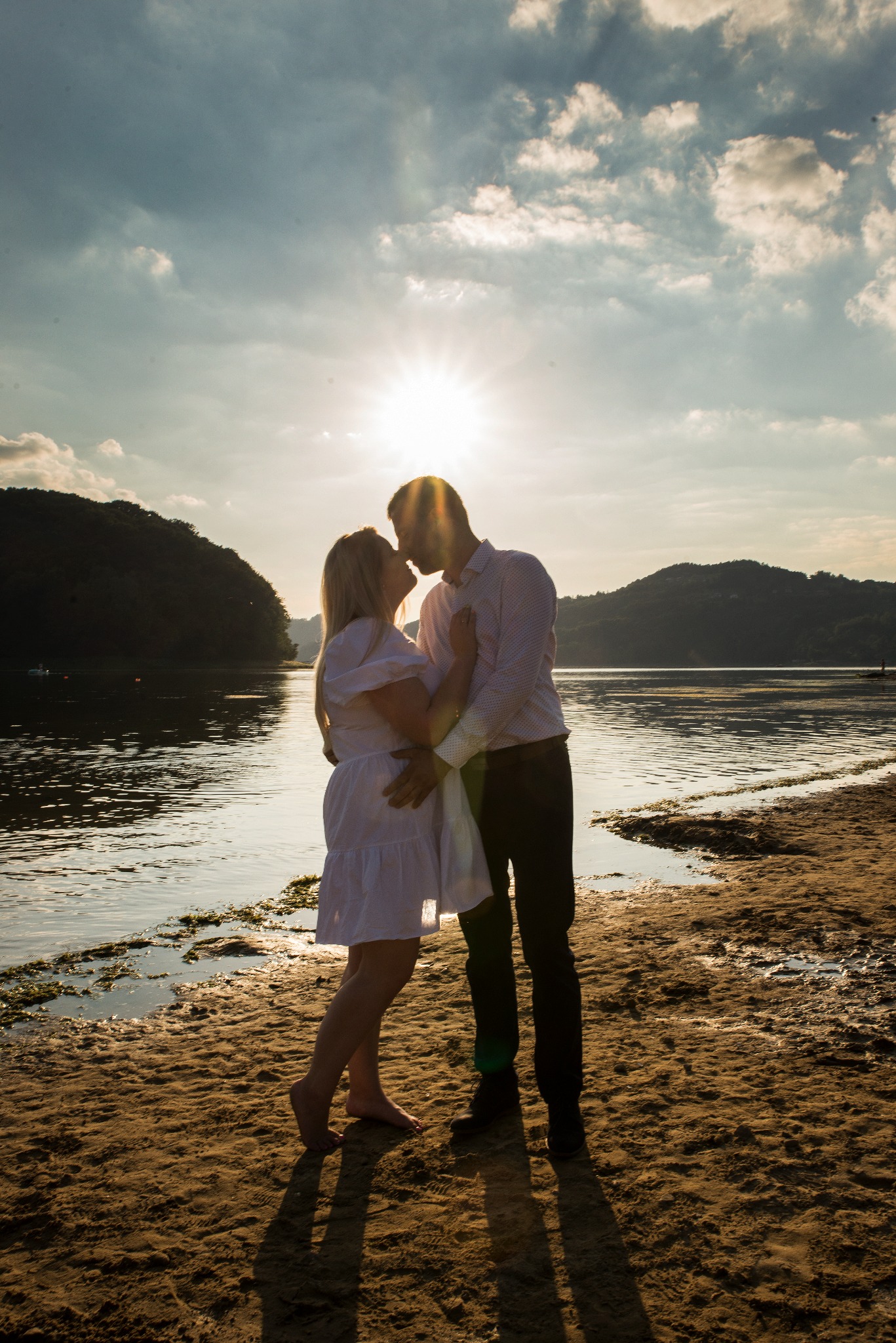 Silhouettes zakochanej pary na plaży w promieniach zachodzącego słońca, kobieta w zwiewnej sukience, mężczyzna w koszuli, romantyczny uścisk na tle wody i zielonych wzgórz.