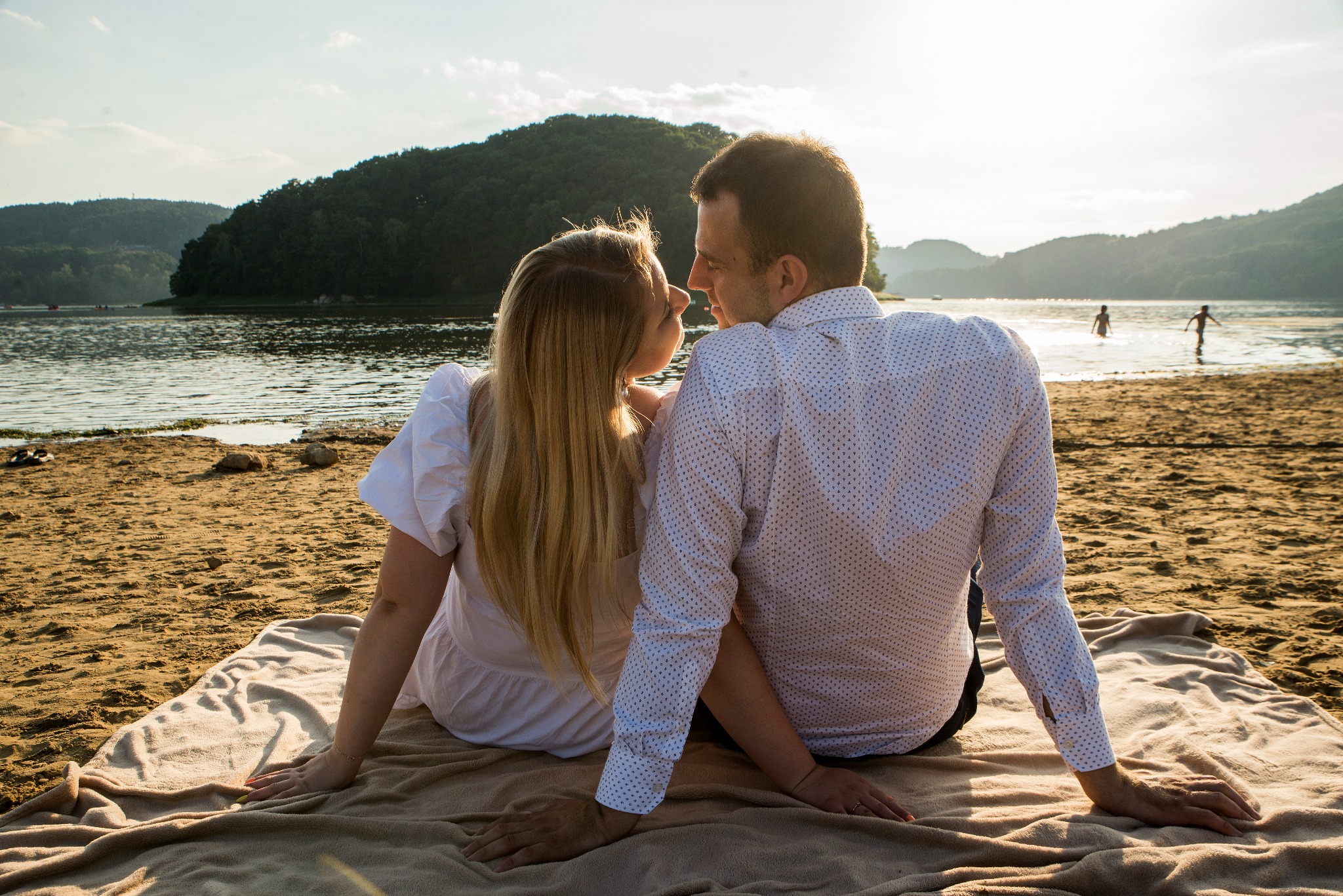 Zakochana para siedzi na kocu na plaży, patrząc na siebie z czułością. W tle woda, góry i sylwetki ludzi w wodzie. Słoneczny dzień.