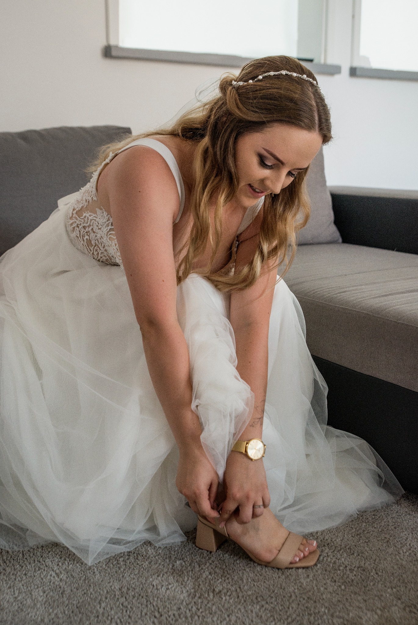 Panna młoda w białej sukni, siedząc na szarej sofie, zapina beżowy but na obcasie. Widoczny złoty zegarek na nadgarstku i delikatna ozdoba we włosach.